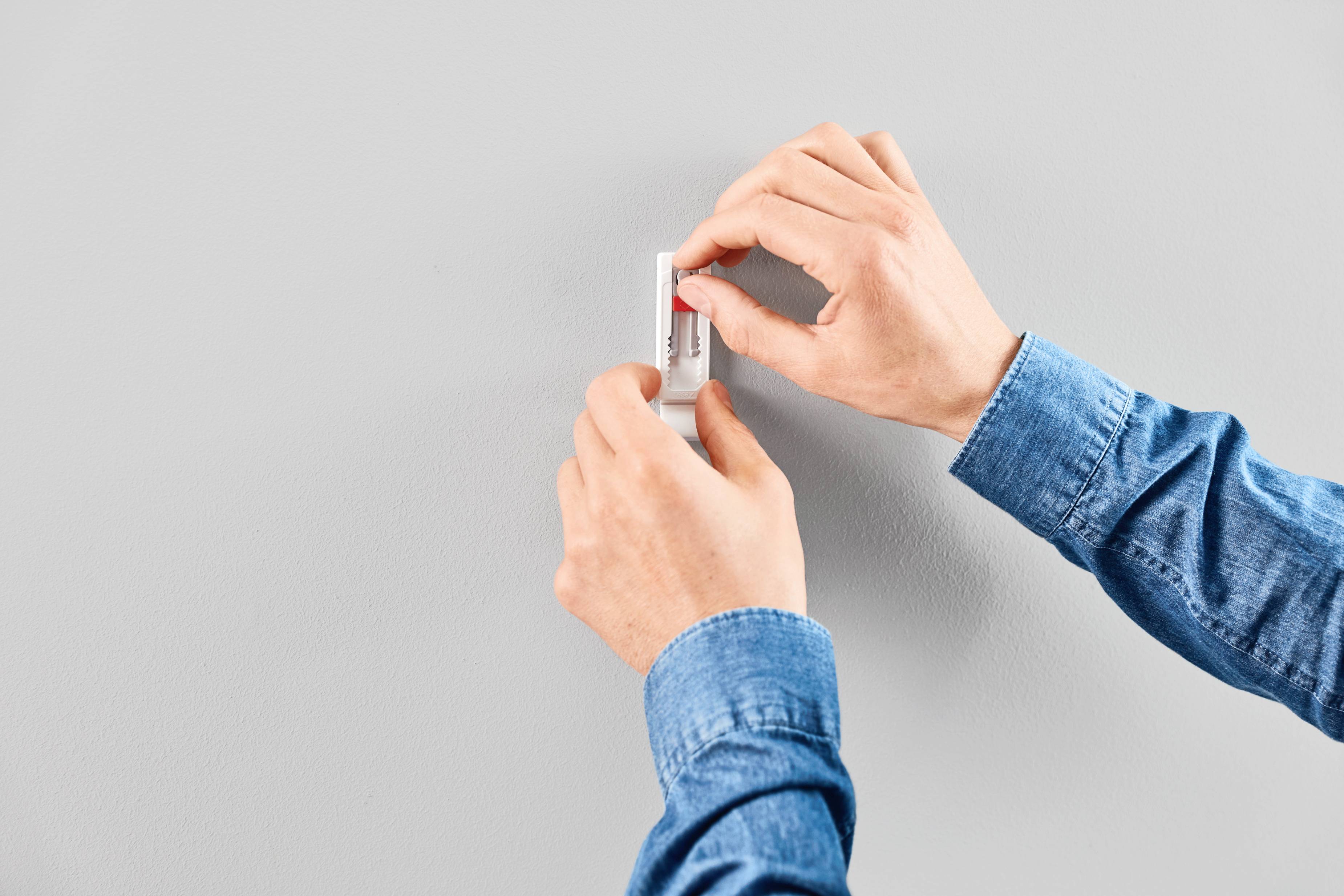 'Person befestigt einen kleinen rechteckigen Gegenstand mit einer klebenden Rückseite an einer grauen Wand.'