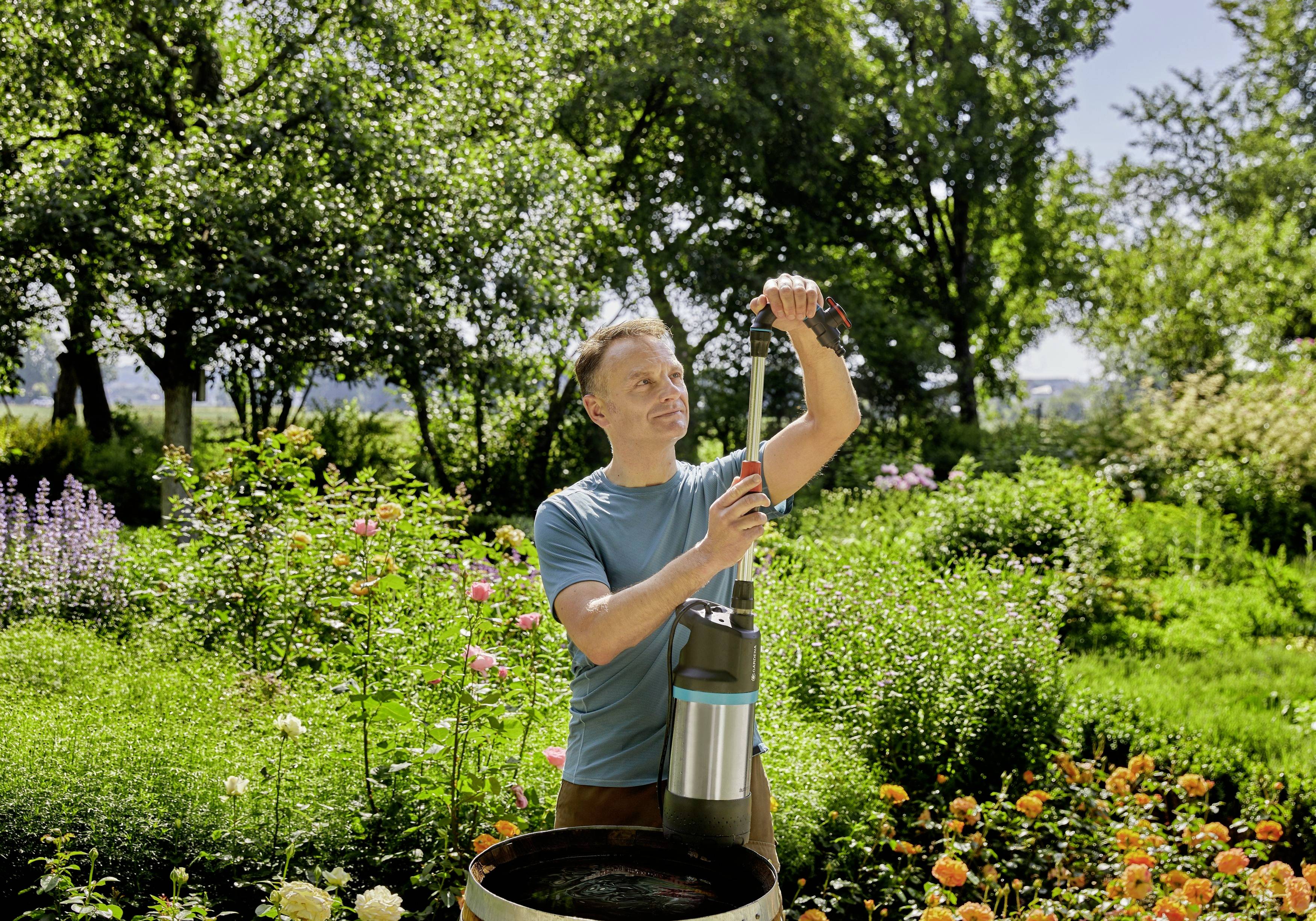 Eine Person verwendet eine Wasserpumpe in einem blühenden Garten. Im Hintergrund sind Blumen und Bäume zu sehen.