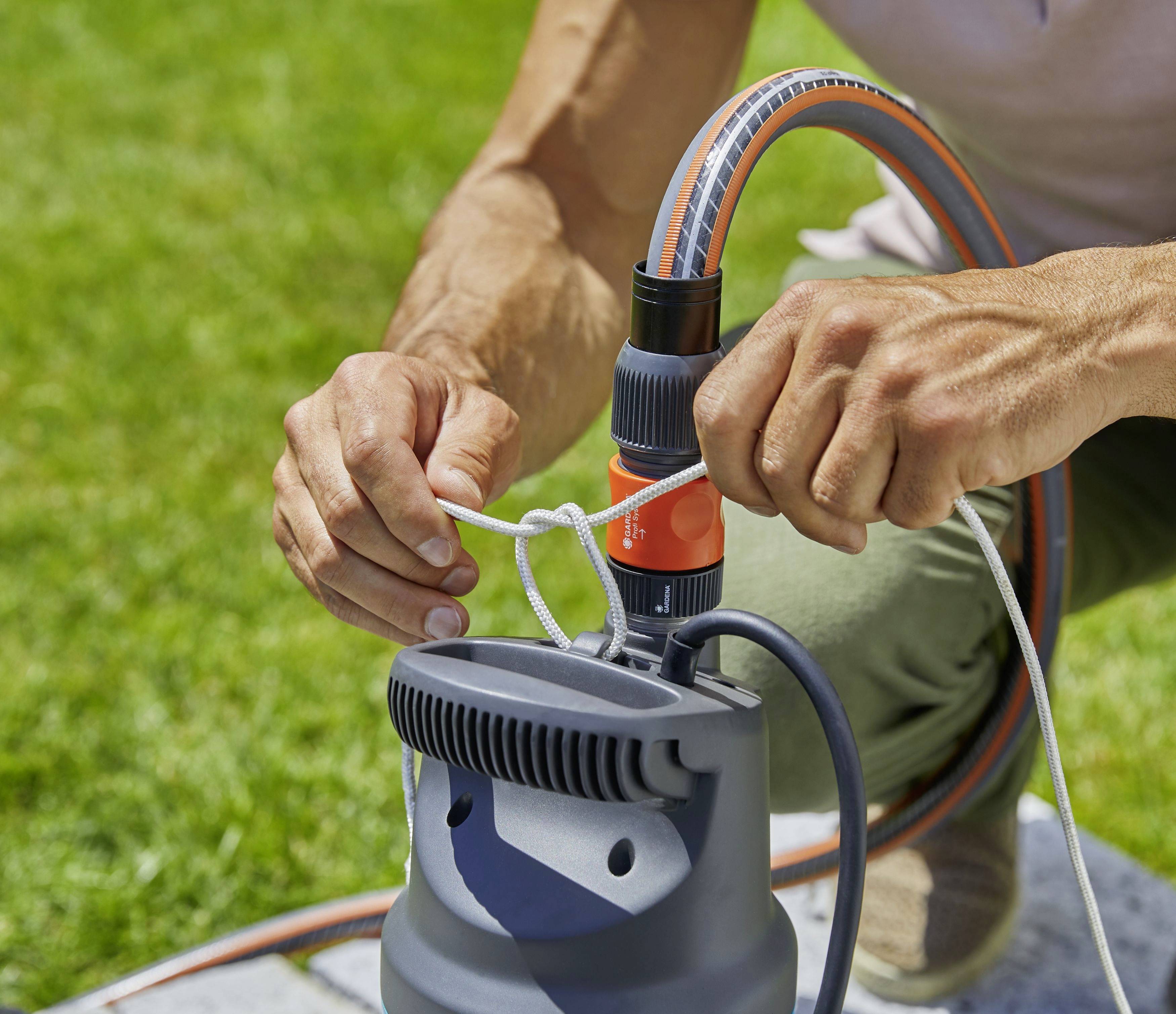 Eine Person bindet ein Seil an eine Pumpe im Freien. Die Pumpe ist mit einem Schlauch verbunden. Rasen im Hintergrund.