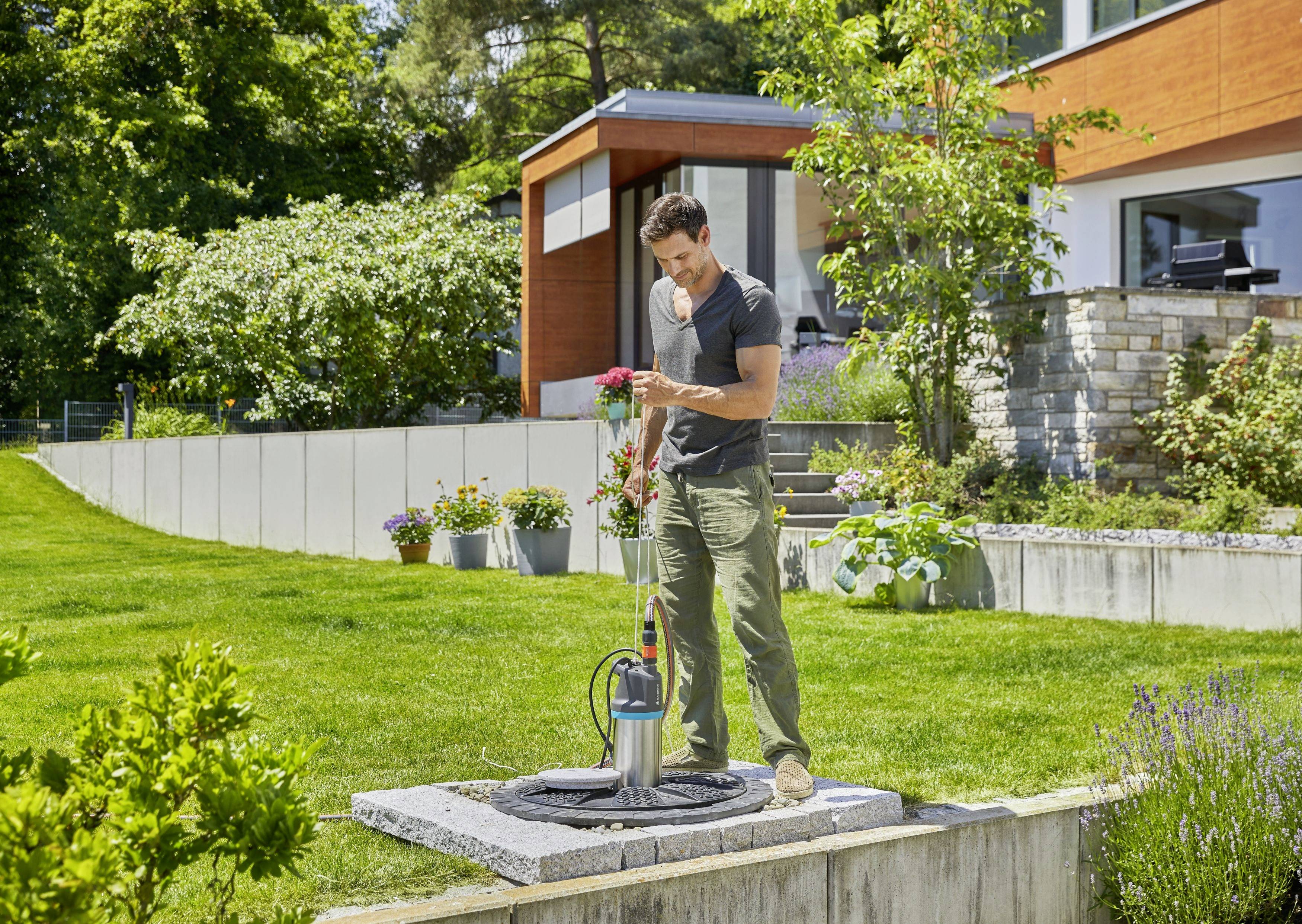 Ein Mann steht in einem Garten neben einem modernen Haus und hält Blumen, während er eine Gartenpumpe überprüft.
