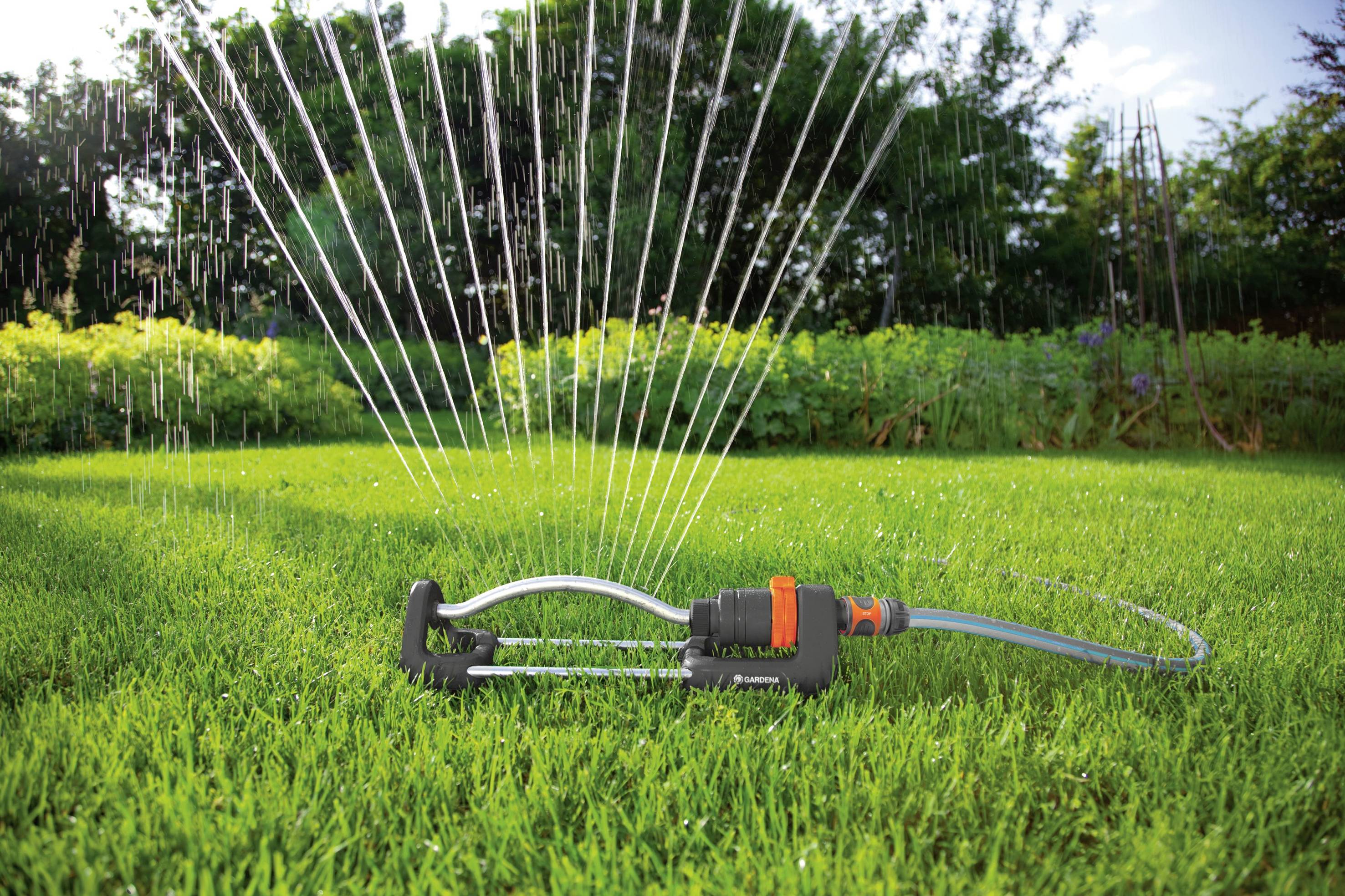 Ein Rasensprinkler bewässert eine grüne Rasenfläche an einem sonnigen Tag. Im Hintergrund sind Bäume und Sträucher sichtbar.