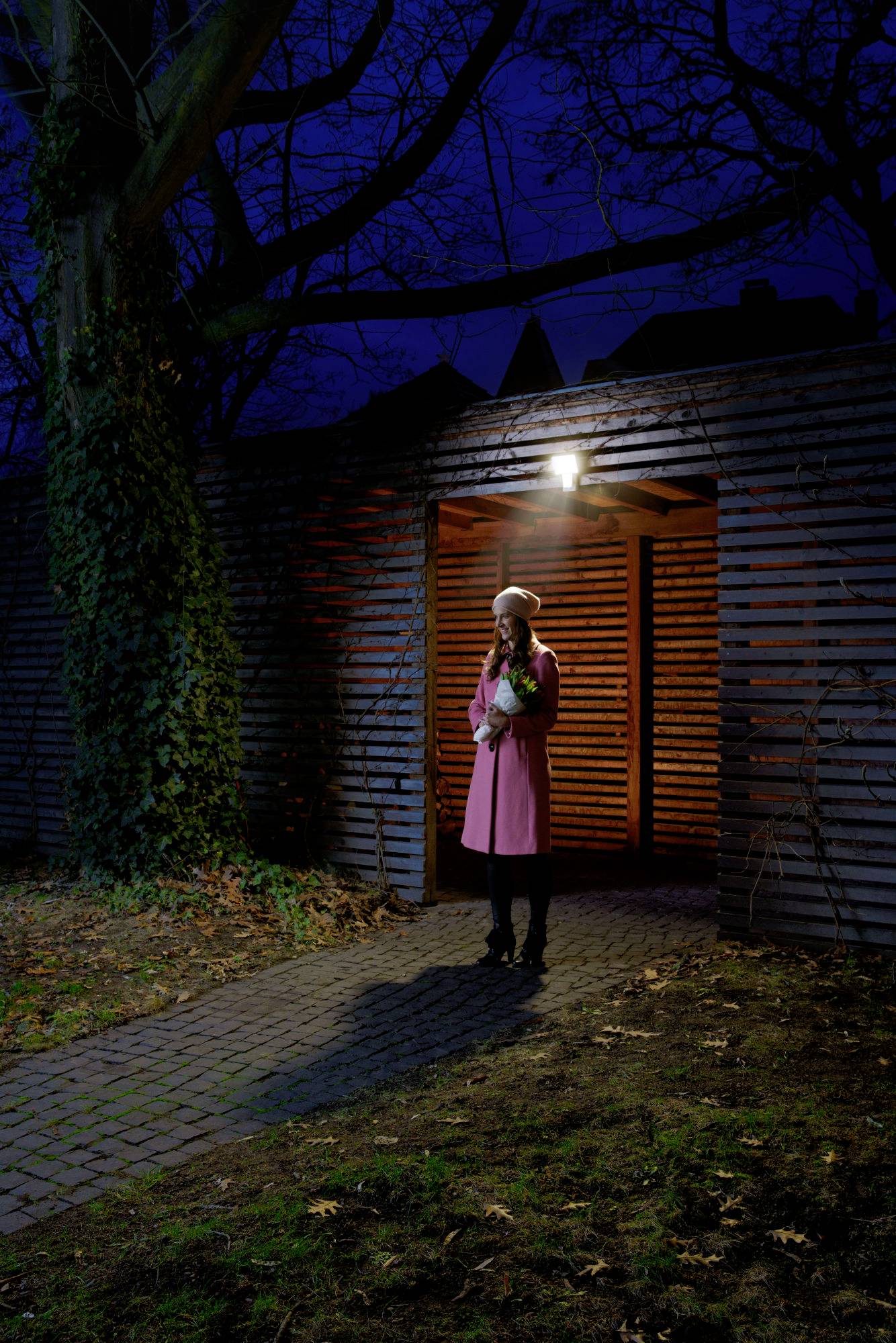 'Frau in rosa Mantel steht abends unter Lampe und hält Blumenstrauß, im Hintergrund Holzwand und Bäume.'