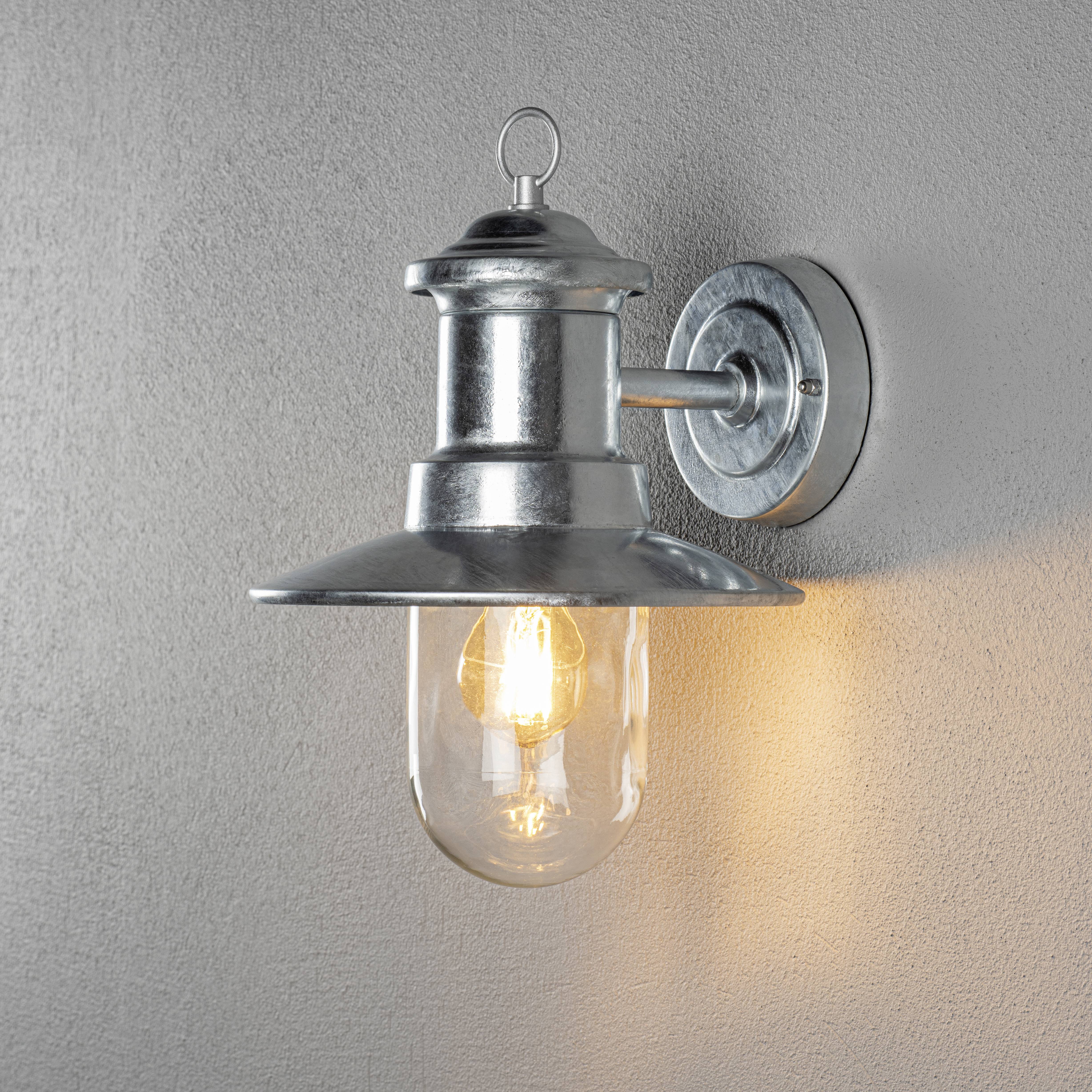 Eine silberne Wandleuchte mit rundem Glasschirm und sichtbarer Glühbirne strahlt warmes Licht auf eine graue Wand.