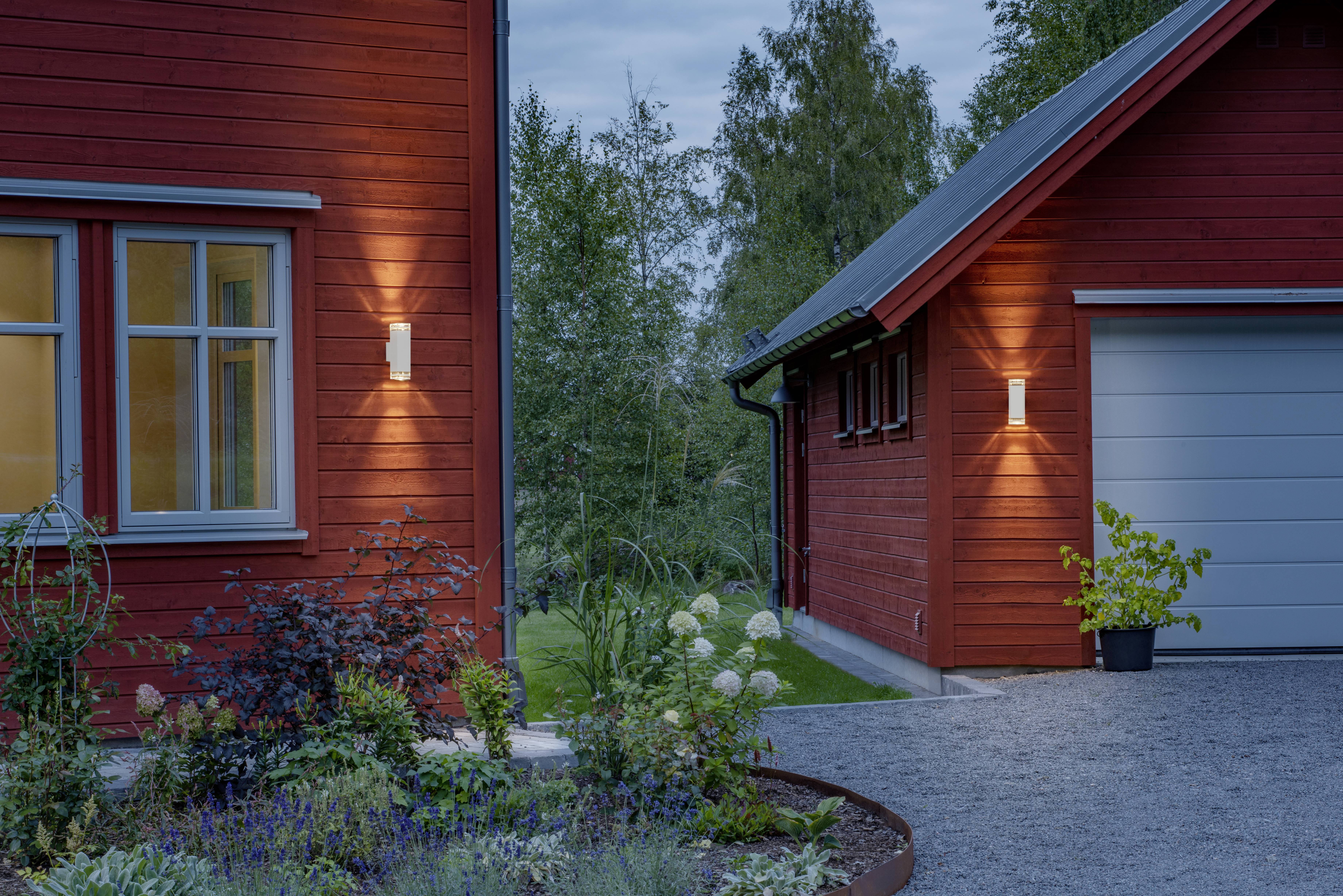 Ein rotes Holzhaus mit eingeschaltetem Außenlicht am Abend, umgeben von einem gepflegten Garten mit Blumen und Sträuchern.