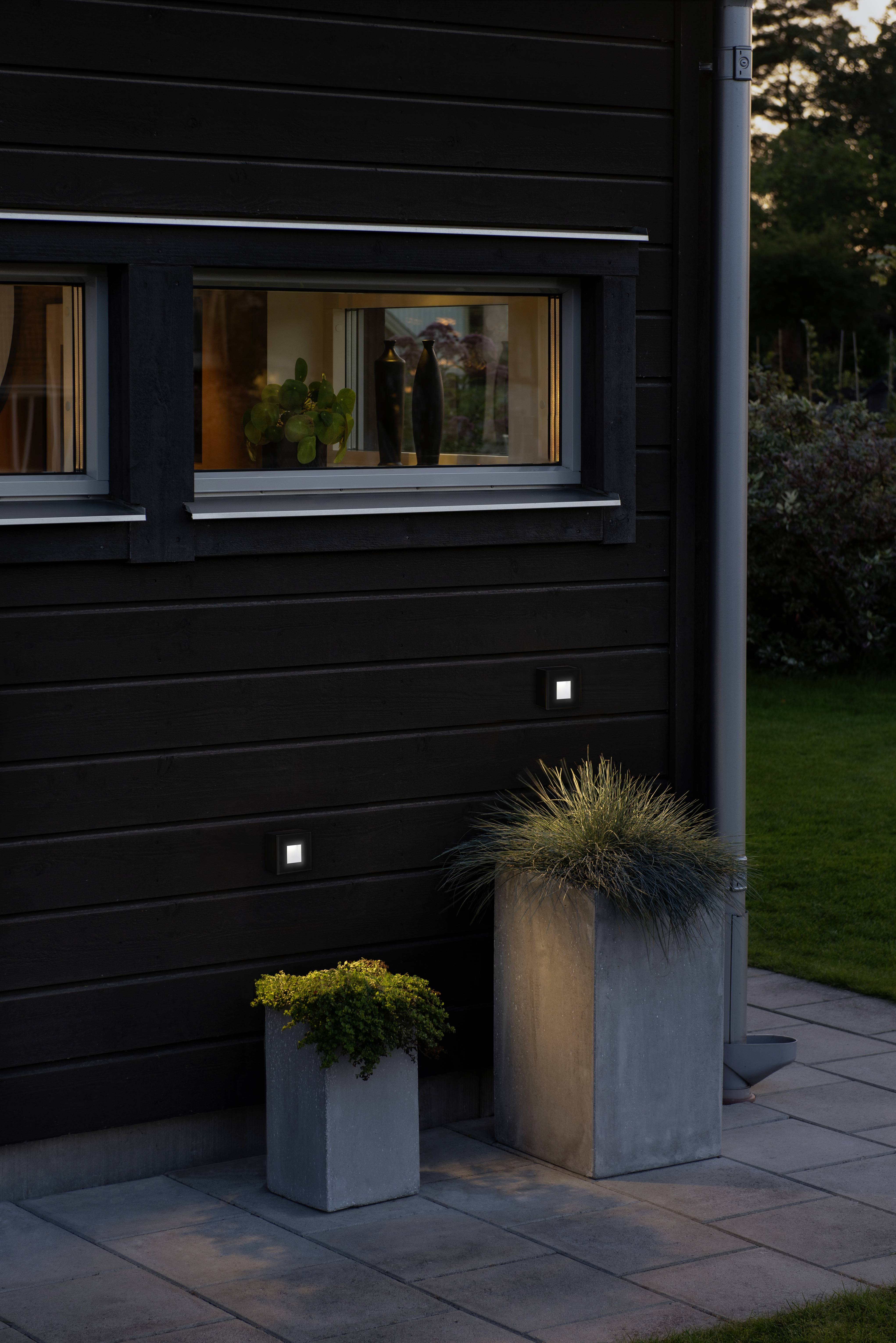 Ein modernes dunkles Haus mit beleuchteten Fenstern in der Abenddämmerung. Zwei Pflanzen in Betonkästen stehen davor auf einer Terrasse.