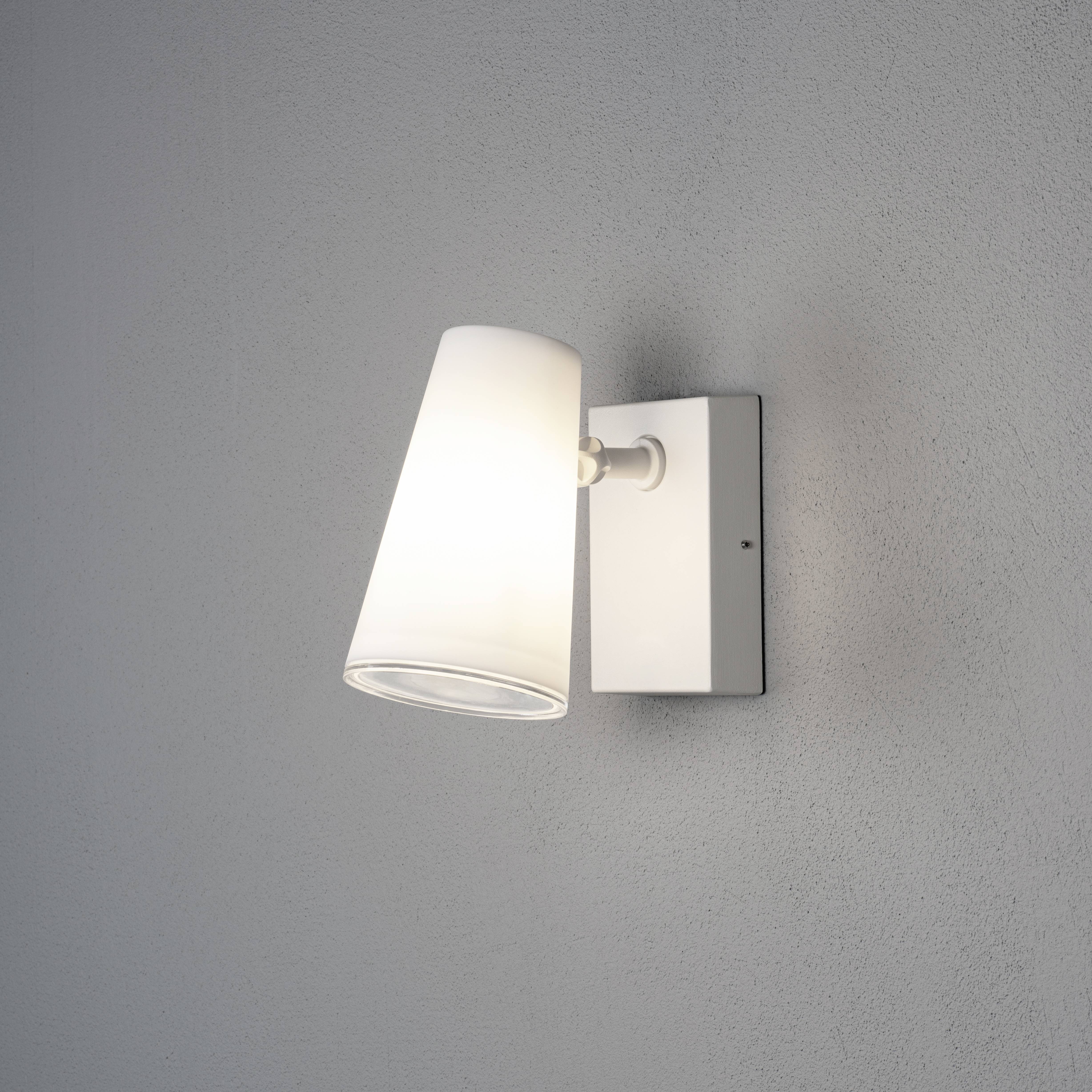 An der grauen Wand hängt eine stilvolle, weiße Wandleuchte mit einem schrägen, zylindrischen Schirm, die ein weiches Licht abstrahlt.