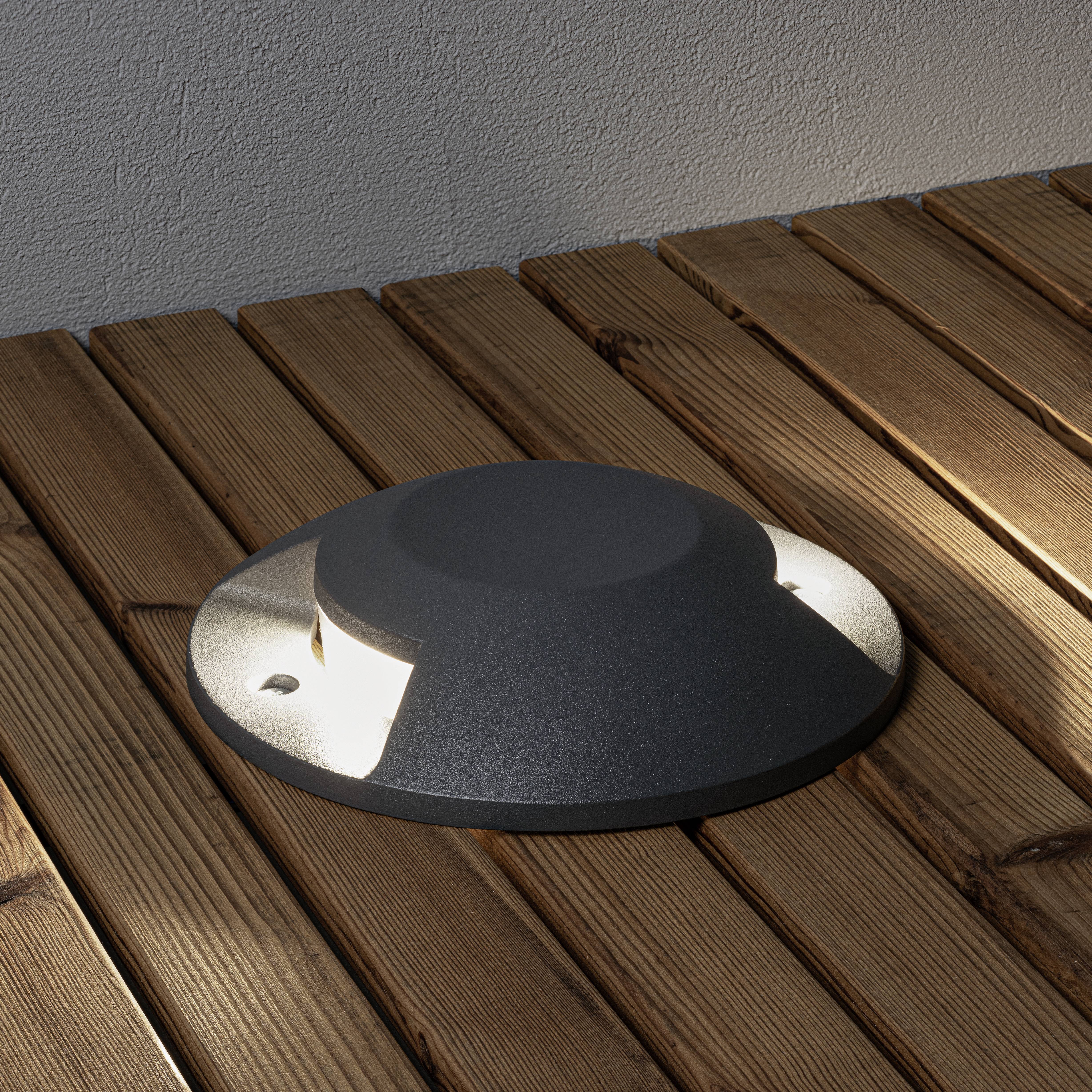 Eine runde, schwarze Bodeneinbauleuchte ist in einem hölzernen Terrassenboden eingebaut. Die Lampe leuchtet und erzeugt ein warmes Licht.