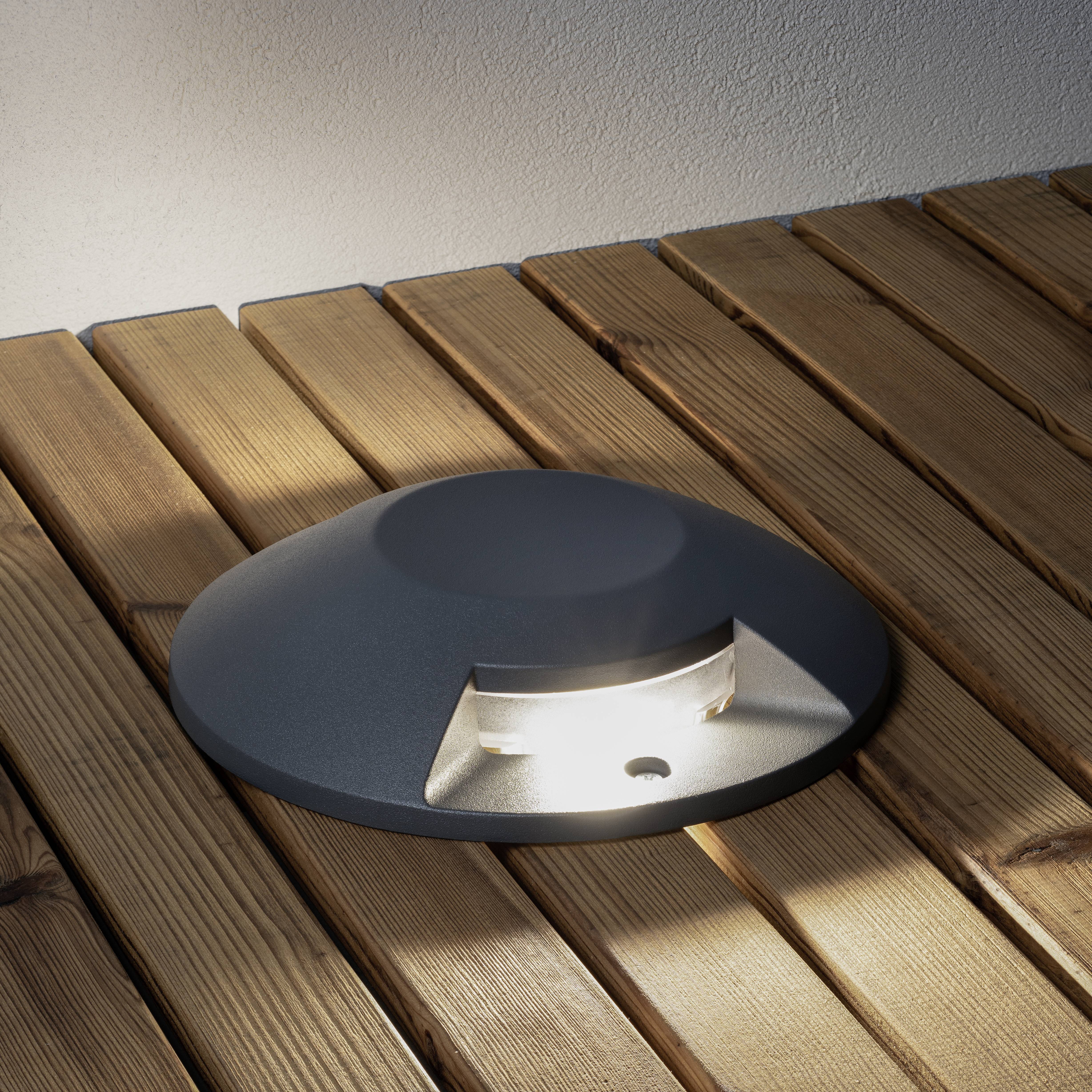 Eine runde Bodenleuchte strahlt warmes Licht auf eine Holzterrasse.