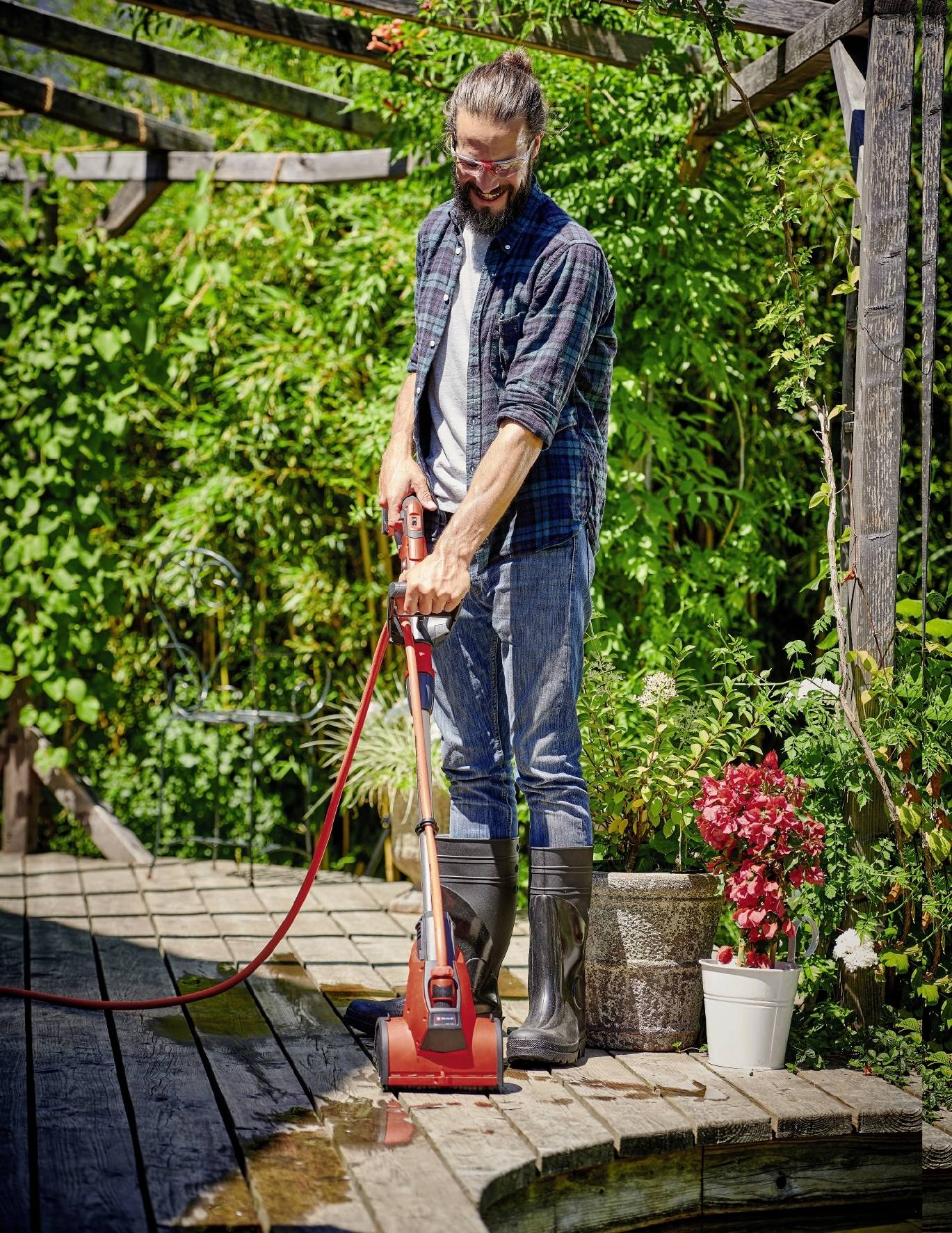 Mann mit langen Haaren, der im Garten mit einer Wasserpumpe auf einer hölzernen Terrasse steht. Umgebung ist grün und blumig.