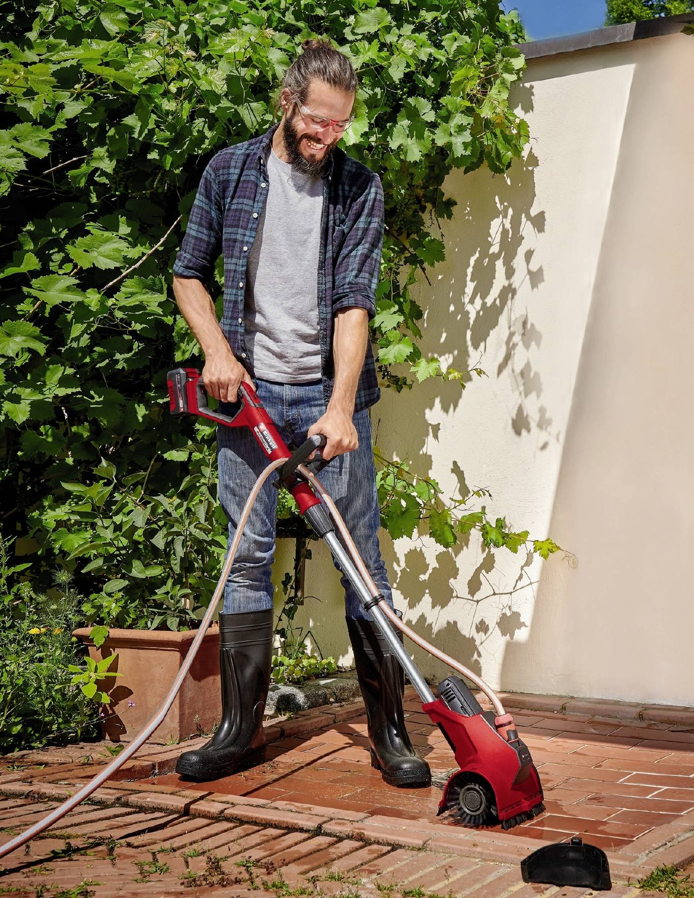 Ein Mann in Gummistiefeln reinigt mit einer rot-schwarzen Maschine die Fugen zwischen Terrassenfliesen. Hintergrund sind Pflanzen und eine weiße Wand.