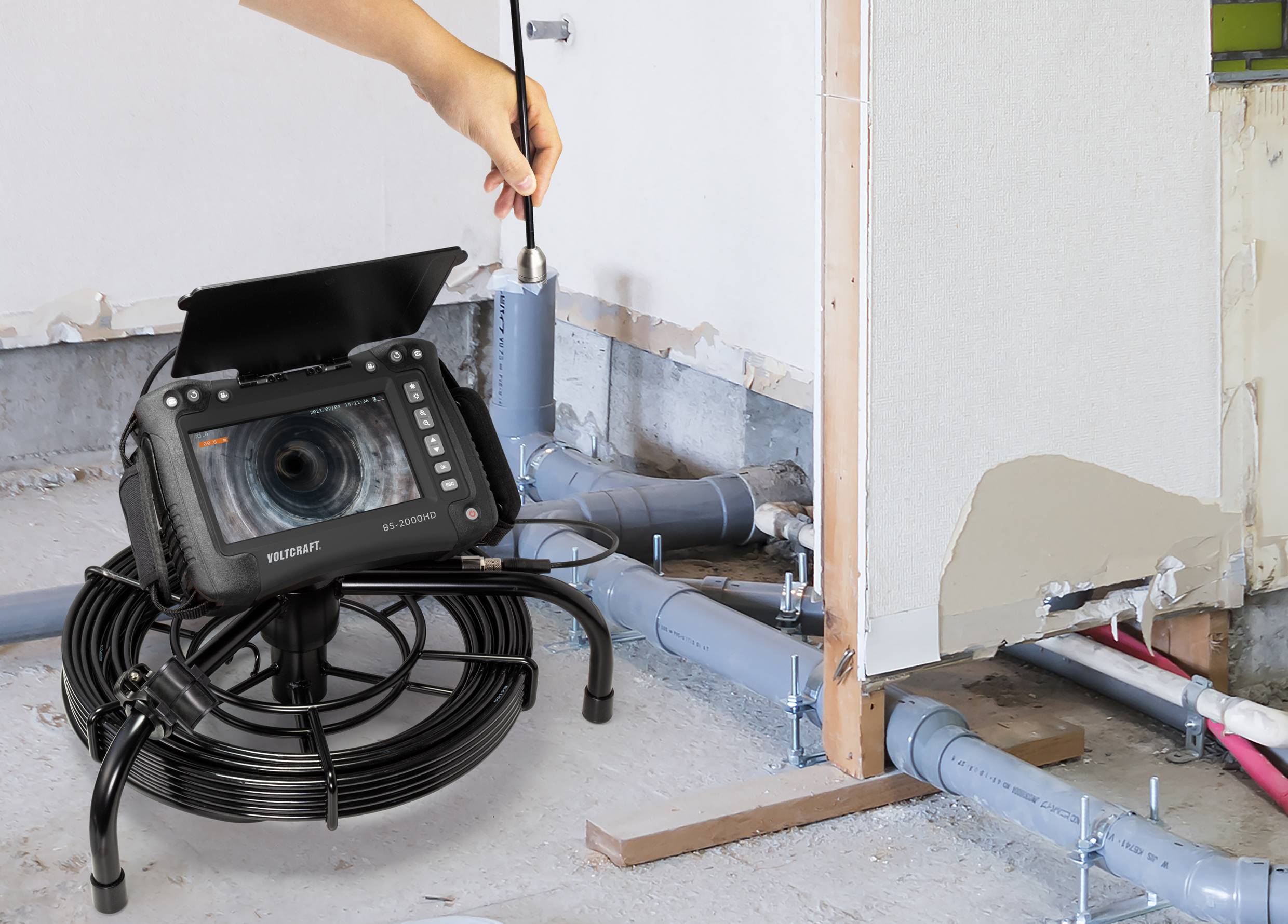 Eine Hand hält eine Inspektionskamera, die auf Rohre im Boden gerichtet ist. Ein Monitor zeigt das Kamerabild, um den Zustand der Rohre zu überprüfen.