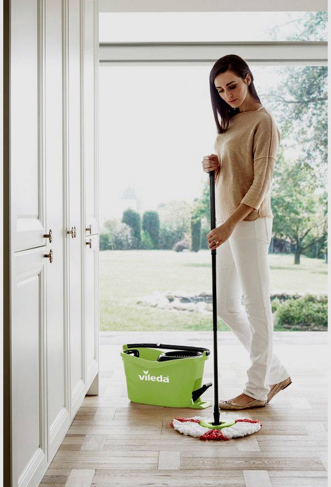 Frau wischt mit einem Mopp den Holzboden in einem hellen Raum. Im Hintergrund steht ein grüner Eimer der Marke 'Vileda'.