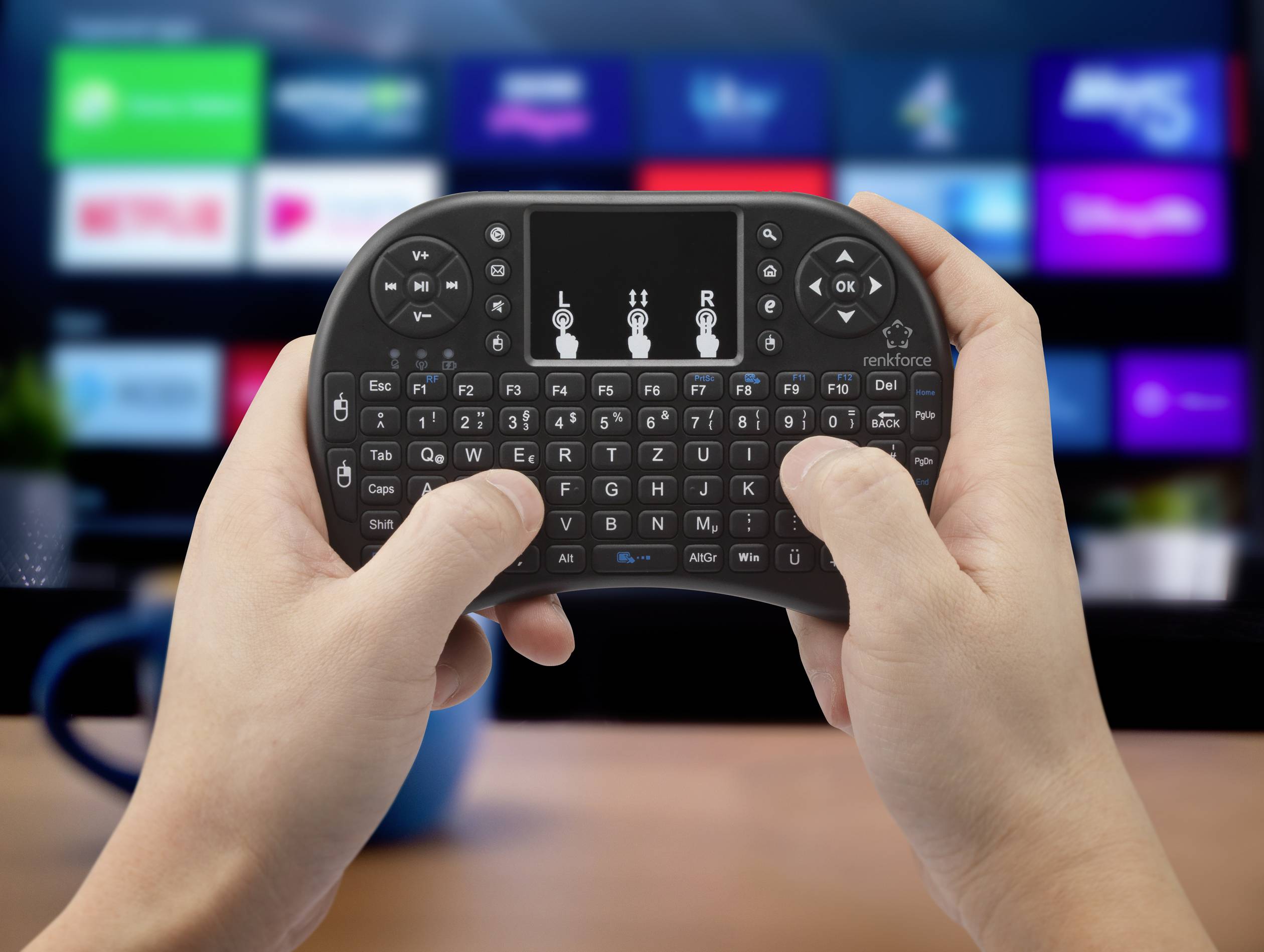 Eine Person hält eine Mini-Tastatur mit Touchpad; im Hintergrund ist ein unscharfer Fernseher mit Streaming-Apps sichtbar.
