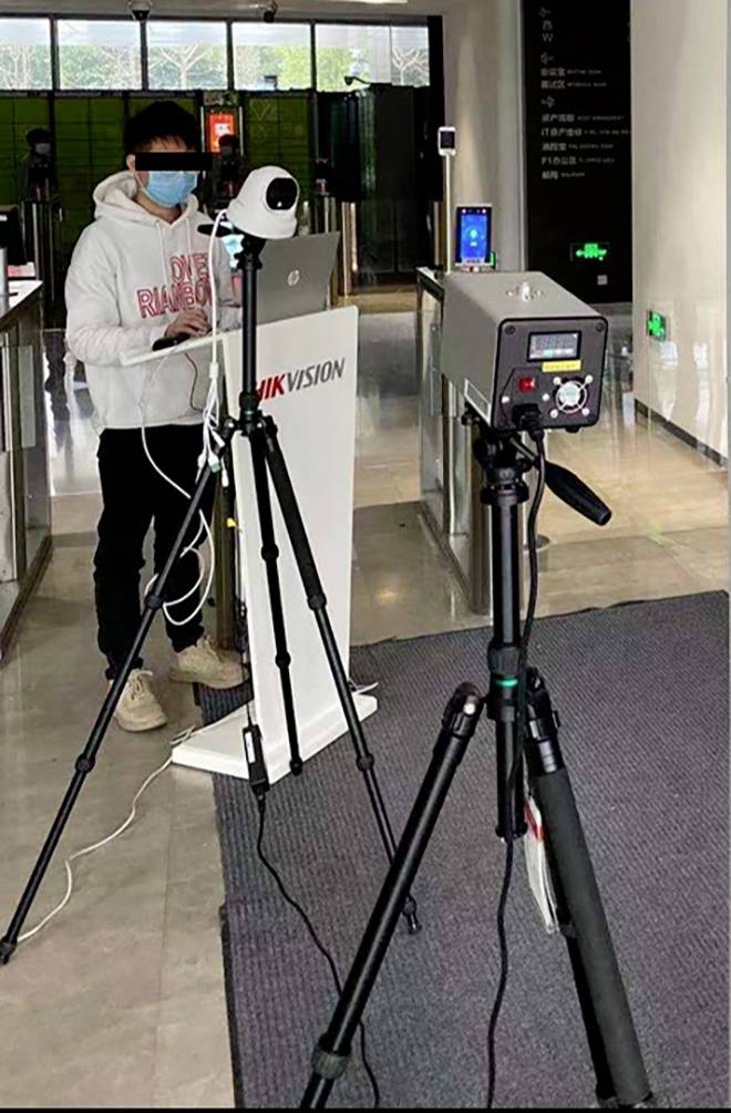 Eine Person mit Maske steht hinter einer Kamera auf einem Stativ in einem Gebäude. Im Hintergrund sind weitere Stative und elektronische Geräte zu sehen.