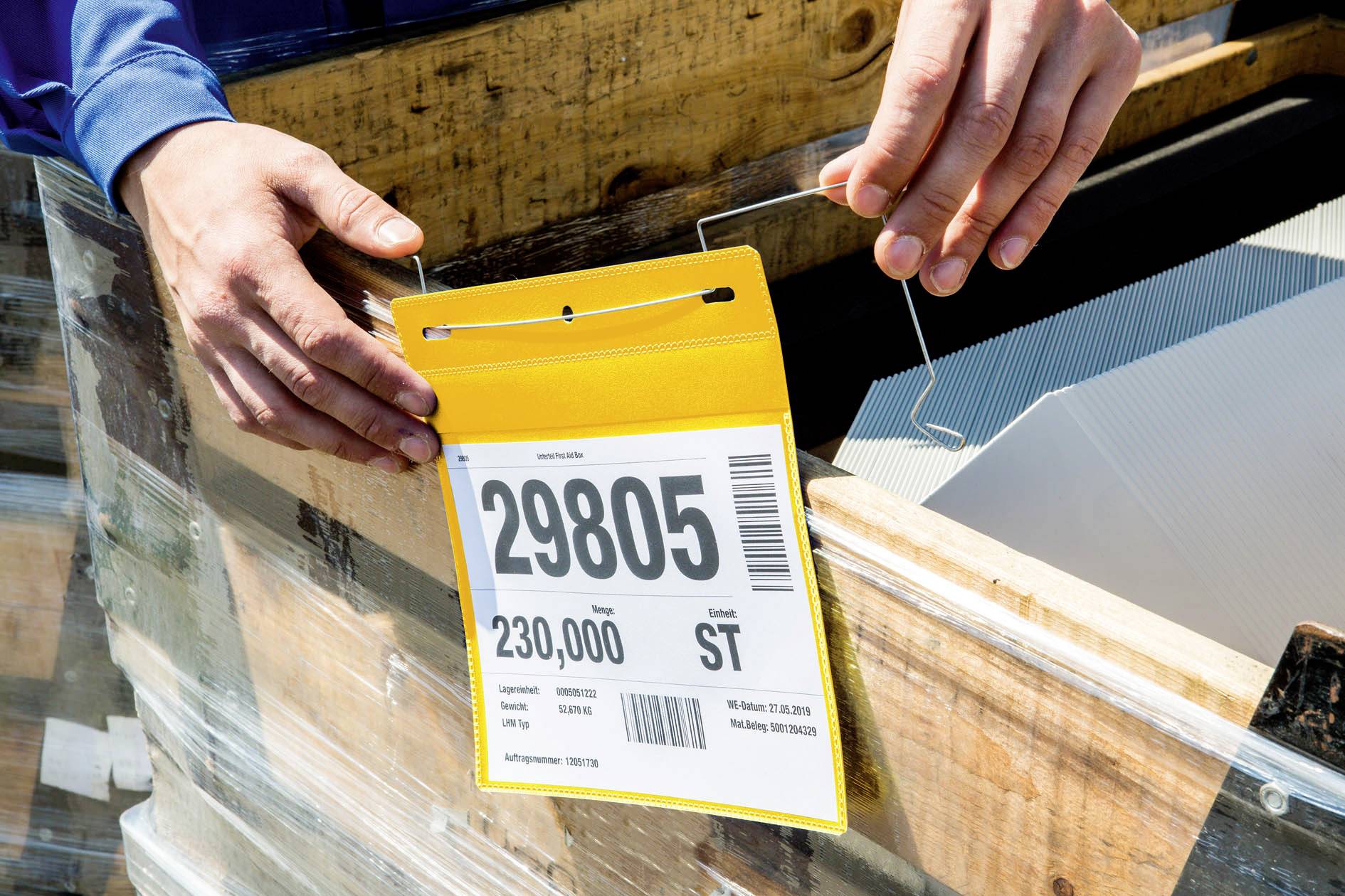 Eine Person hält ein Schild mit der Aufschrift '29805' und '230.000 St'. Es ist an einer Holzpalette mit verpackten Materialien befestigt.