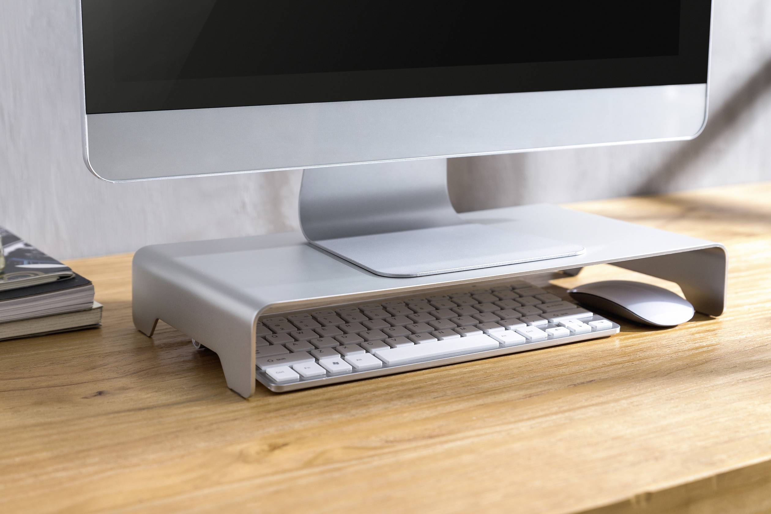 Ein Computermonitor steht auf einem erhöhten Ständer aus Metall. Darunter befinden sich eine Tastatur und eine Maus auf einem Holztisch.