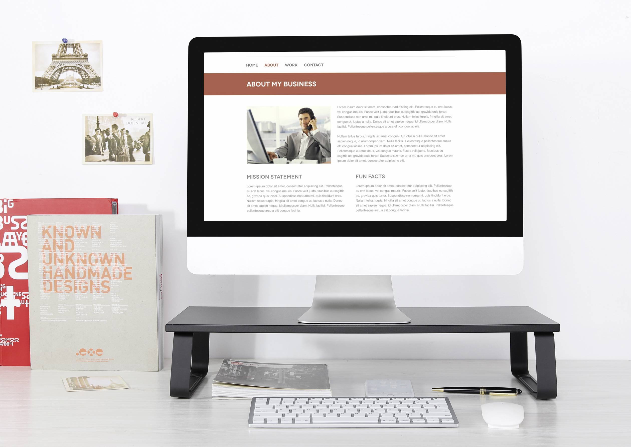 Ein Desktop-Computer mit einer Webseite, die 'About My Business' zeigt, steht auf einem Tisch. Daneben sind Bücher und ein Poster.