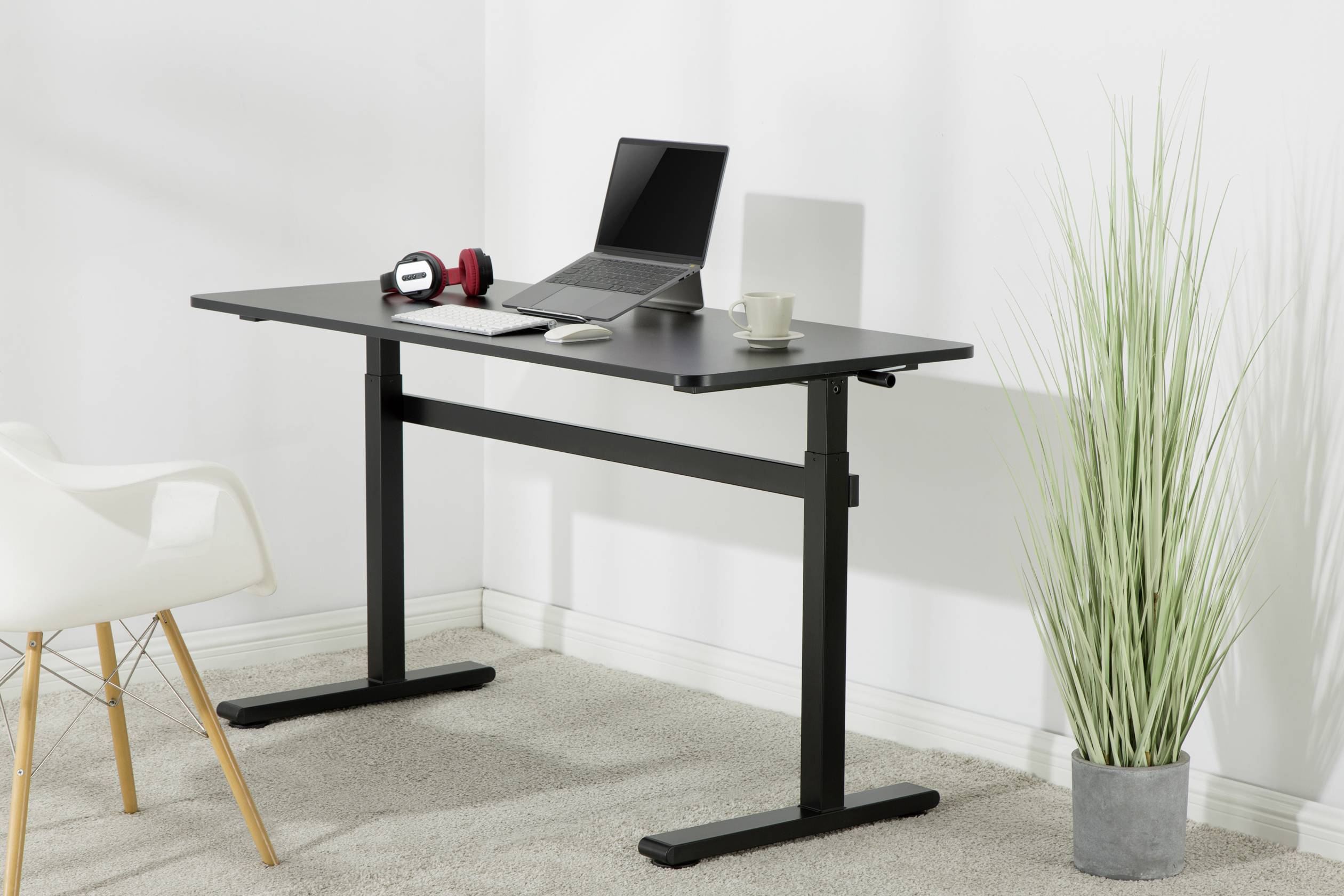 Ein höhenverstellbarer Schreibtisch mit einem Laptop, Kopfhörern und einer Tasse, daneben steht eine grüne Pflanze auf dem Boden.