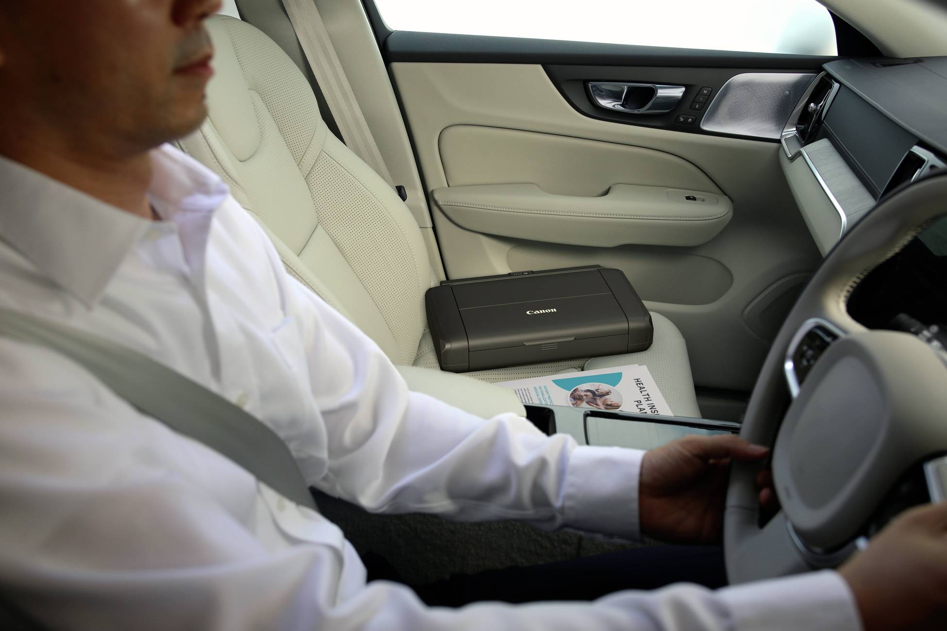 Ein Mann im Auto steuert das Fahrzeug. Ein mobiler Drucker und ein Magazin liegen auf dem Beifahrersitz.