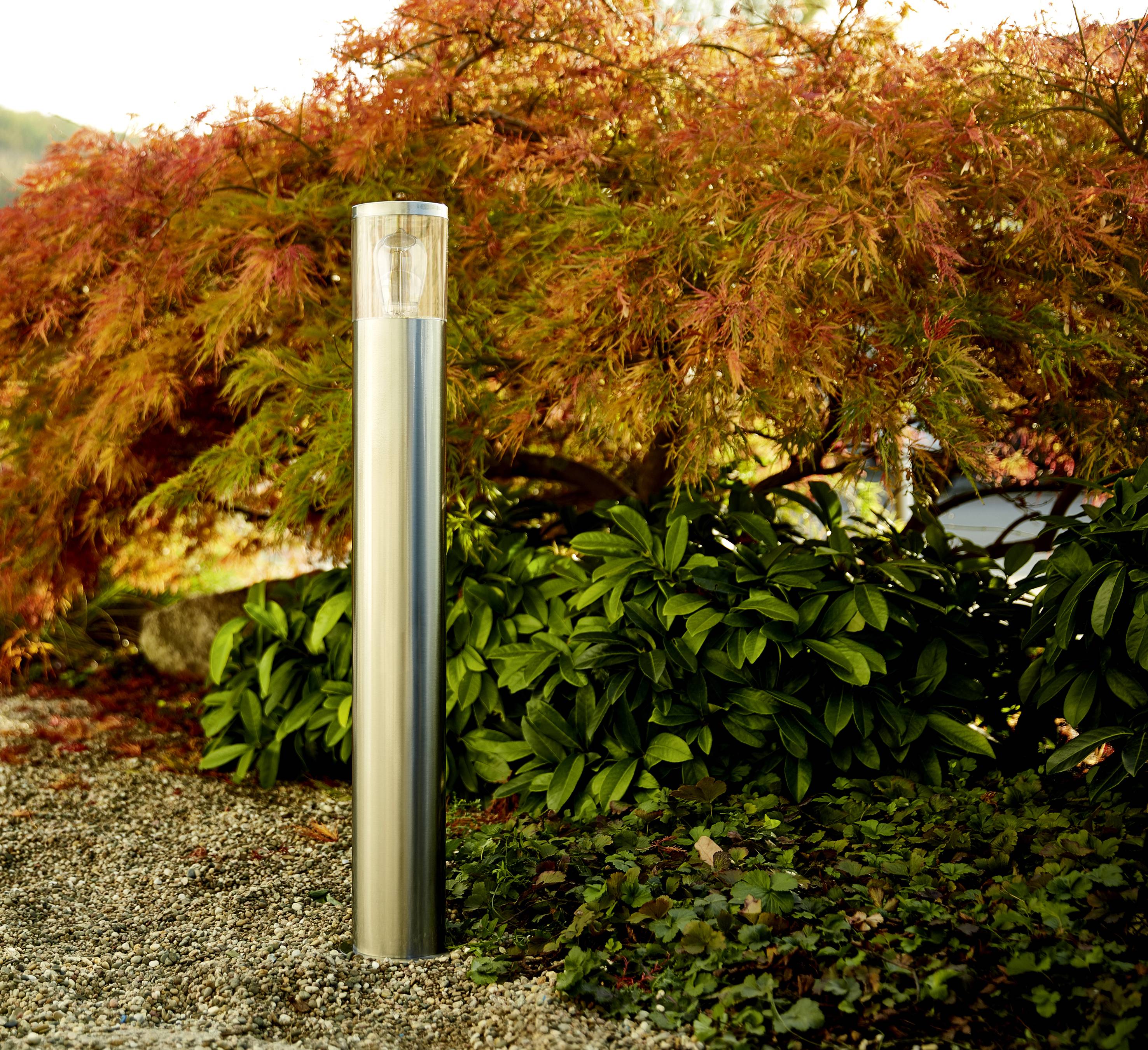 Eine moderne Gartenlaterne im Vordergrund, umgeben von grünem Laub und einem Baum mit leuchtend roten Blättern im Hintergrund.