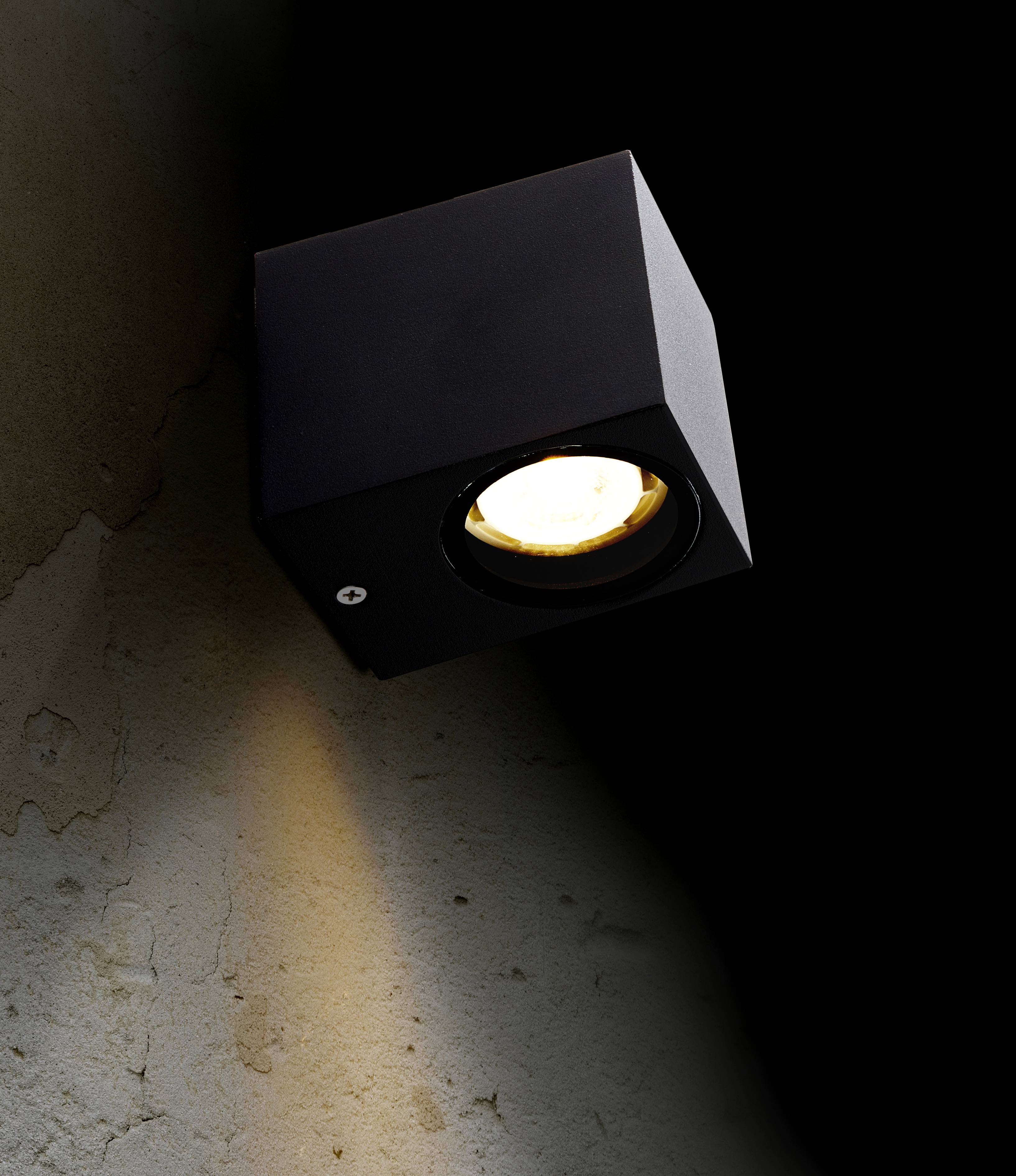 Eine schwarze Wandlampe in Würfelform leuchtet an einer leicht strukturierten, beige-grauen Wand und erzeugt einen warmen Lichtstrahl nach unten.