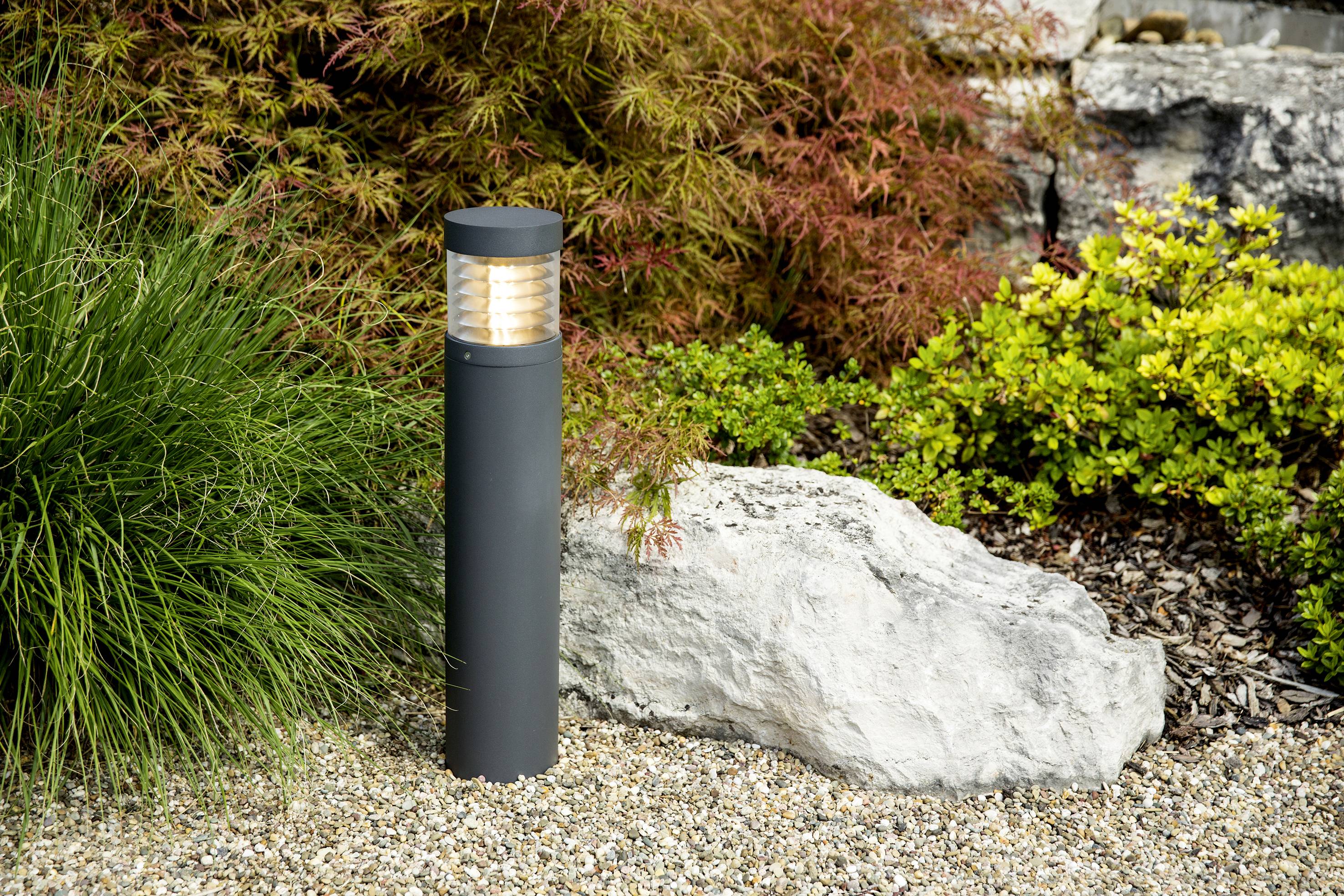 Eine moderne Gartenlampe leuchtet auf einem kiesbedeckten Weg neben grünen und roten Pflanzen; ein großer Stein im Hintergrund.