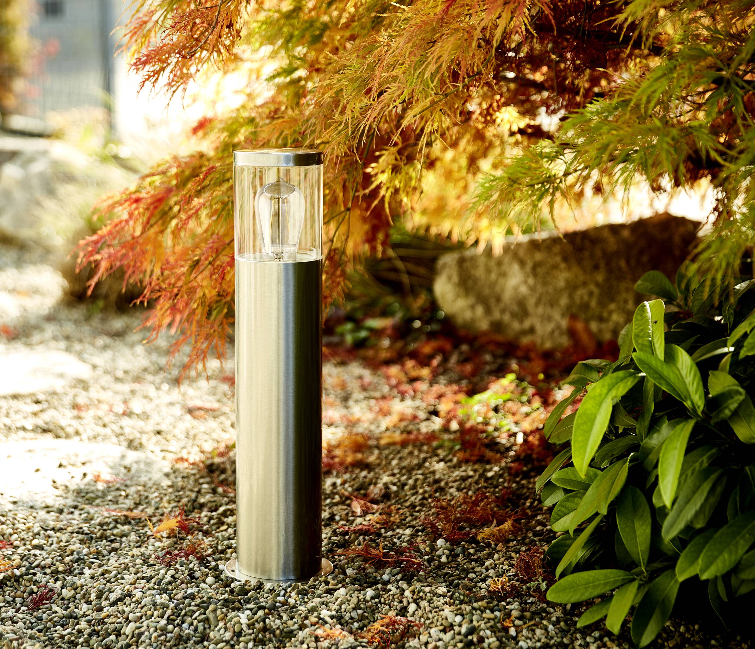 Eine moderne Außenlampe aus Metall steht auf einem Kiesweg, umgeben von herbstlichen Pflanzen mit roten und grünen Blättern.