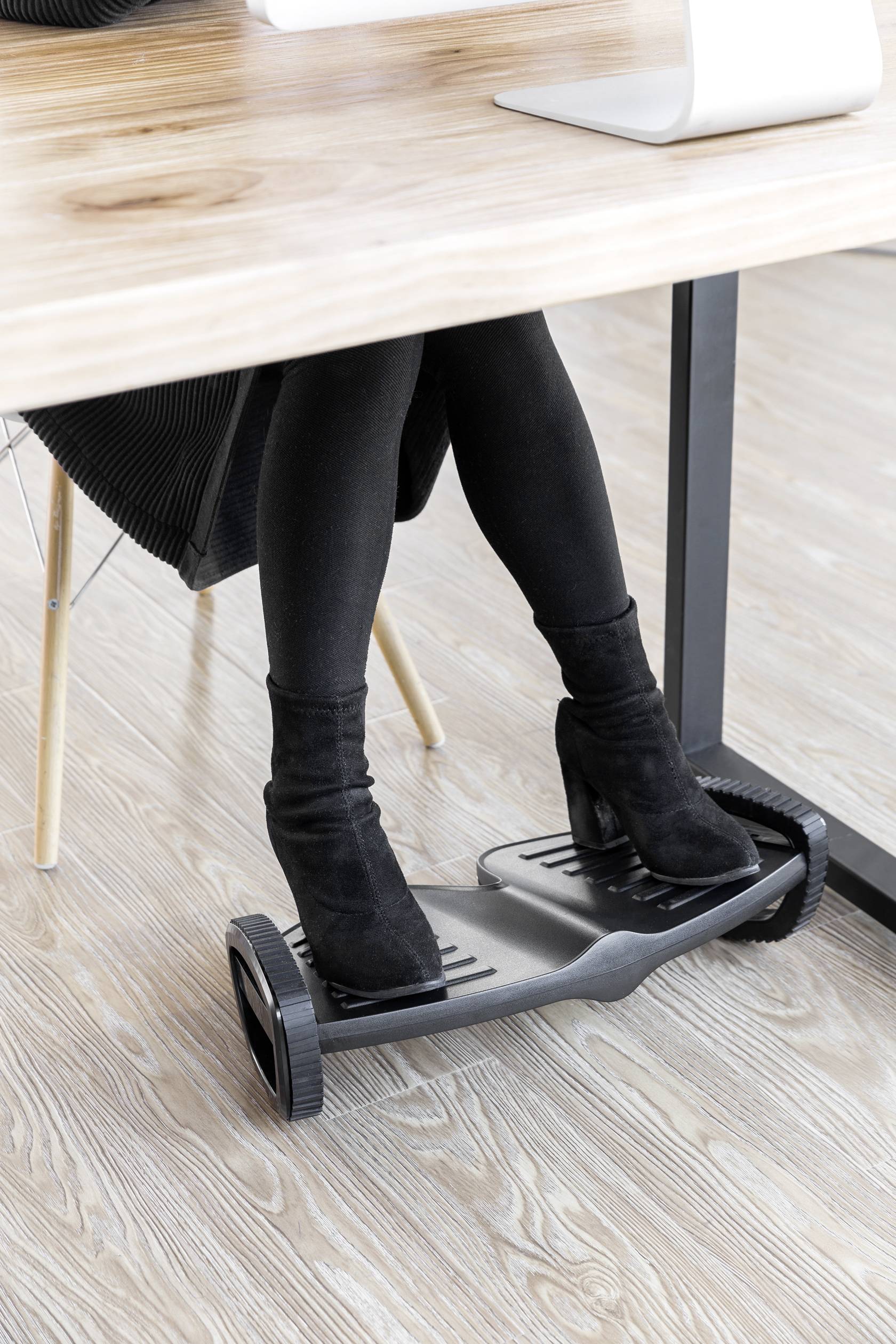 Eine Person sitzt an einem Schreibtisch und benutzt eine Fußstütze mit Rollen. Die Person trägt schwarze Stiefel und schwarze Kleidung.