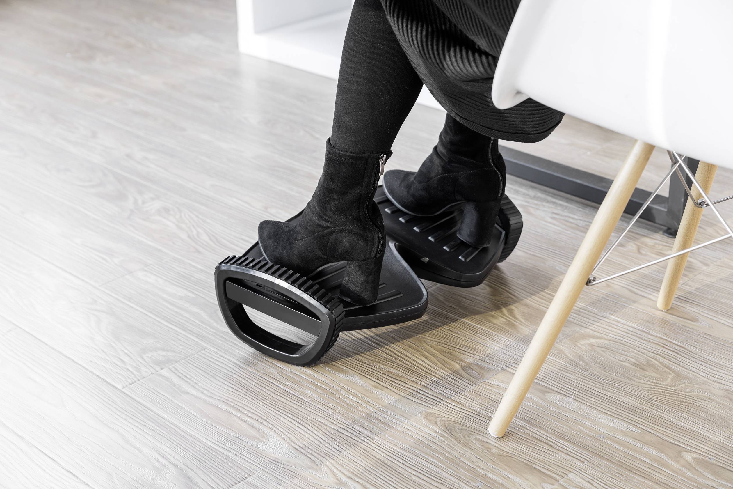 Beine einer Person, die schwarze Stiefel trägt, ruhen auf einer schwarzen Fußstütze unter einem Schreibtisch auf einem Holzboden.