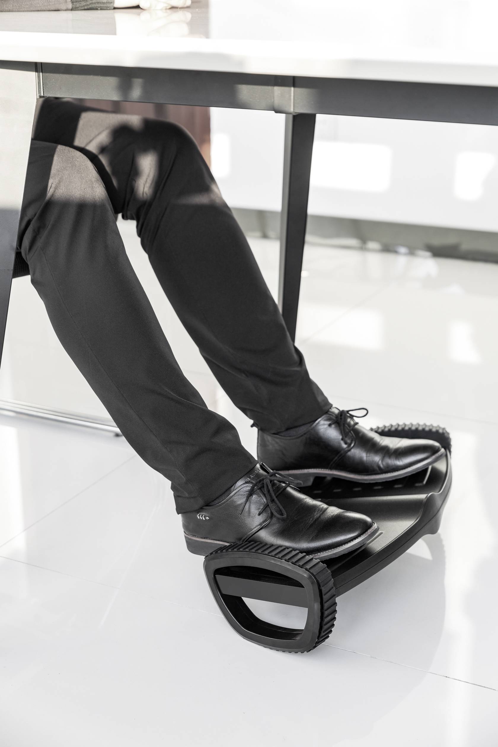 Eine Person in schwarzen Hosen und Schuhen ruht ihre Füße auf einem schwarzen Fußhocker unter einem Tisch.