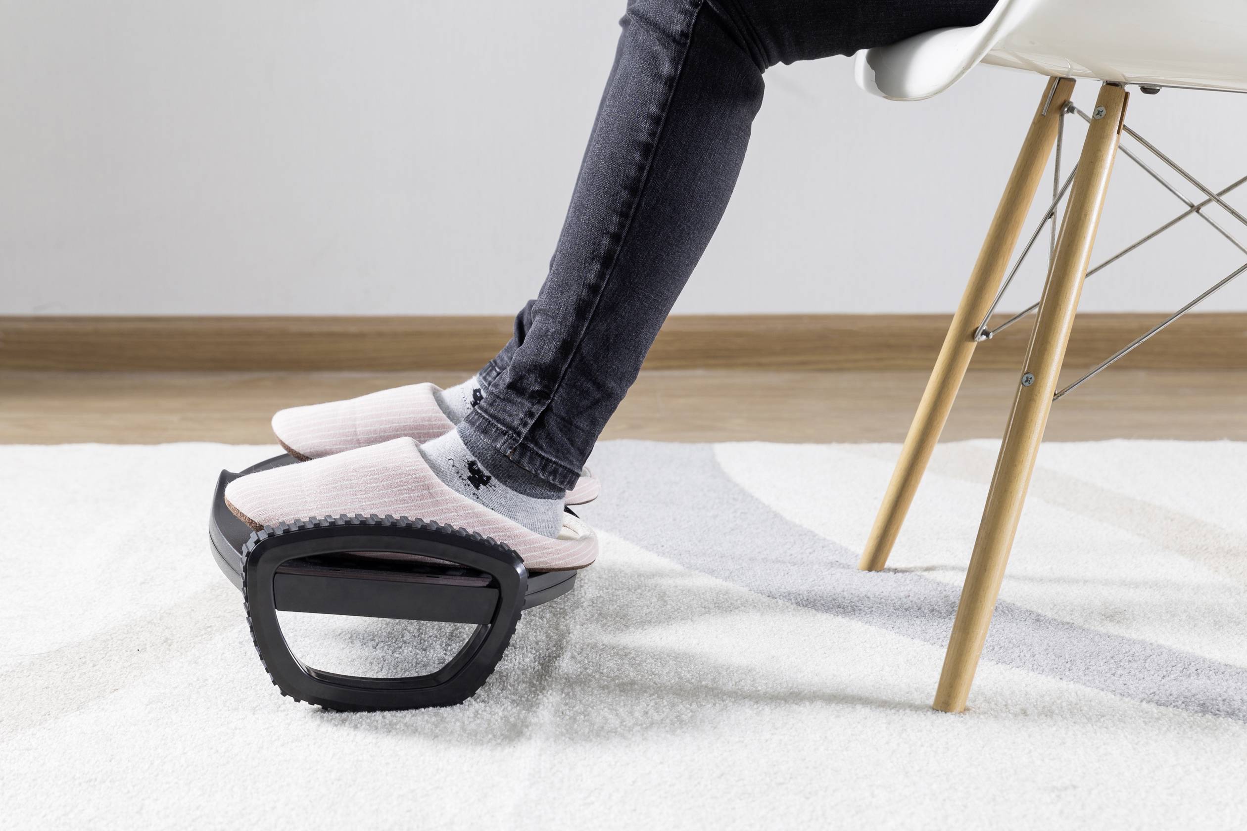 Eine Person sitzt auf einem Stuhl und ruht ihre Füße in Hausschuhen auf einem Fußstützenroller. Teppich und Holzboden im Hintergrund.