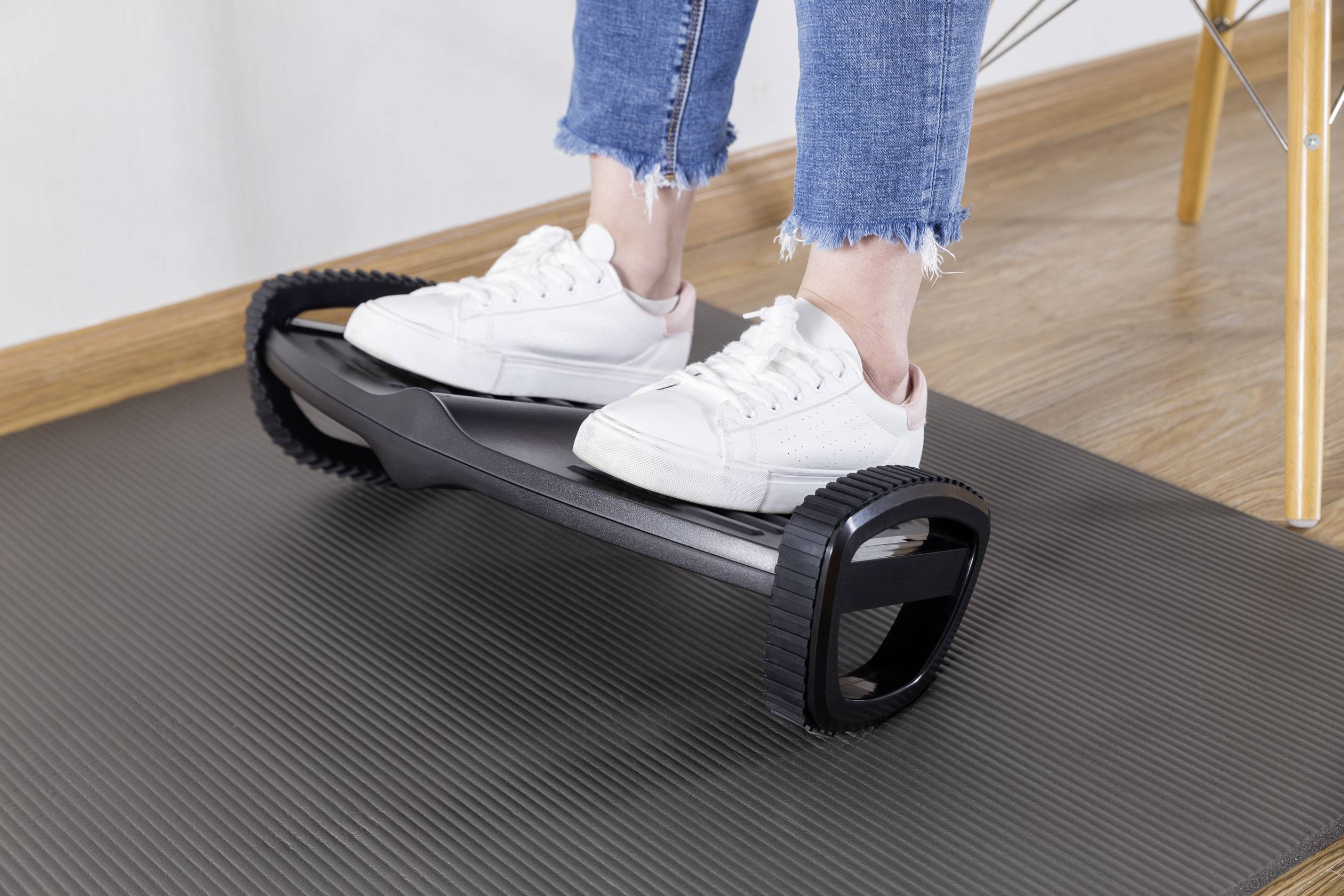 Eine Person balanciert auf einem Balance-Board mit weißen Turnschuhen. Das Board liegt auf einer dunklen Matte auf dem Boden.