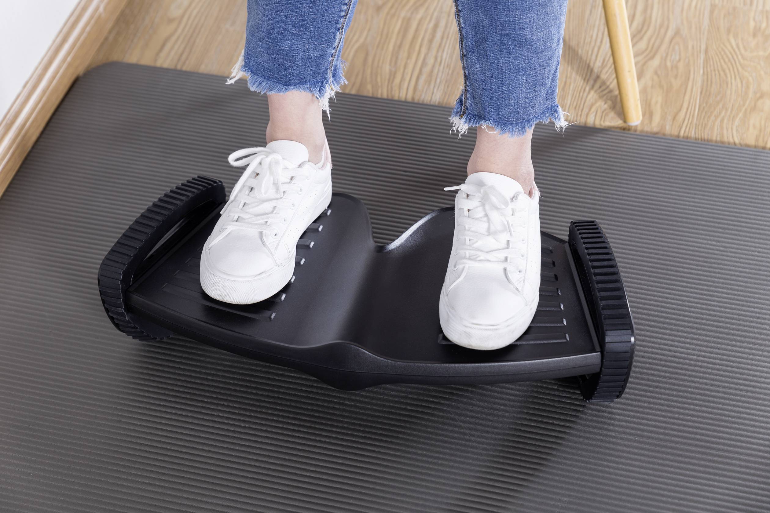 Eine Person steht auf einem Balance-Board mit schwarz gerillter Oberfläche, trägt weiße Turnschuhe und blaue Jeans auf einem Holzboden.