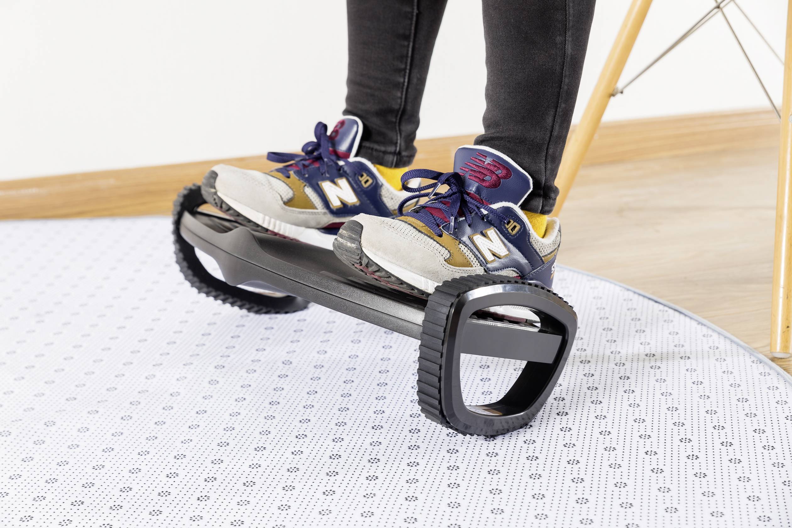 Person steht auf einem einradartigen Balancegerät mit zwei großen Reifen. Sie trägt Sportschuhe. Der Boden ist mit einem gemusterten Teppich bedeckt.