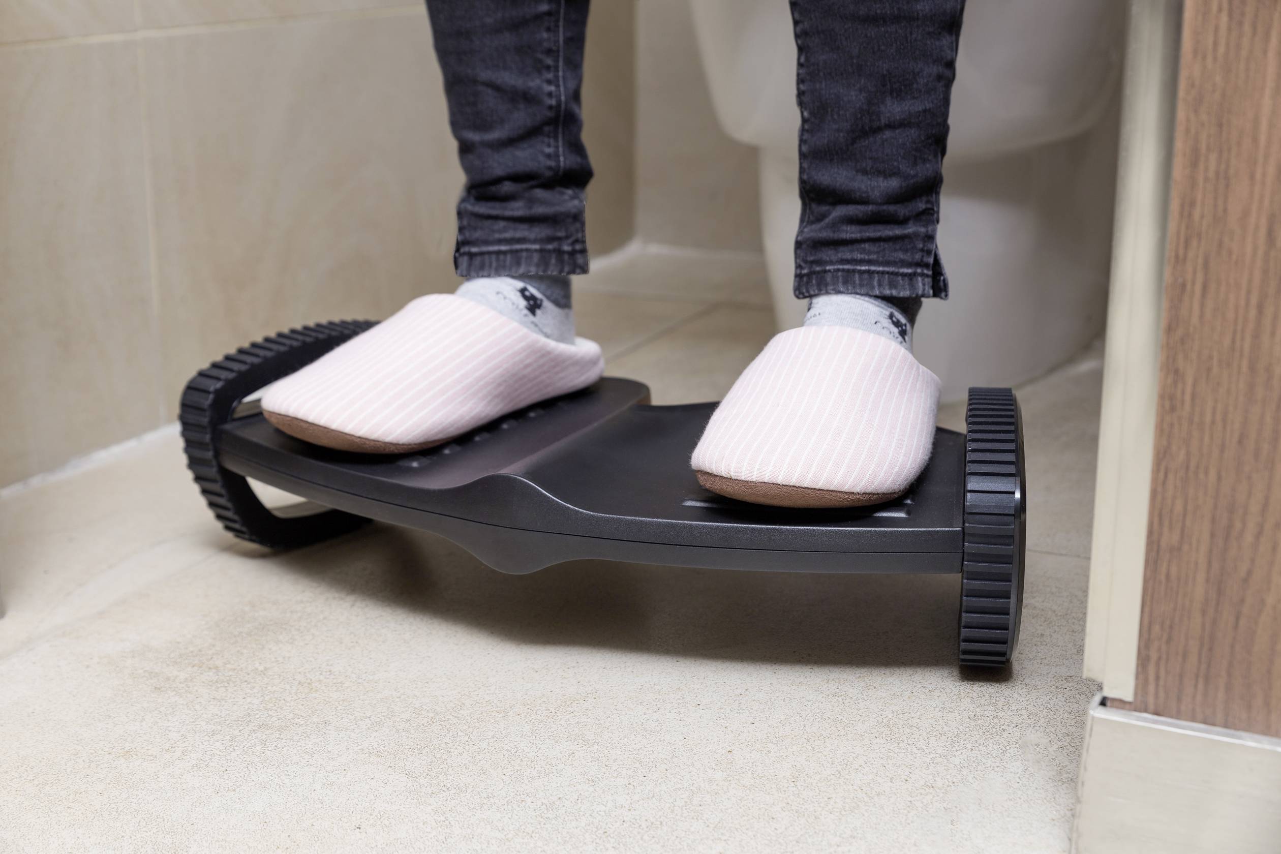 Füße in rosa Hausschuhen stehen auf einer höhenverstellbaren Fußstütze neben einer Toilette.