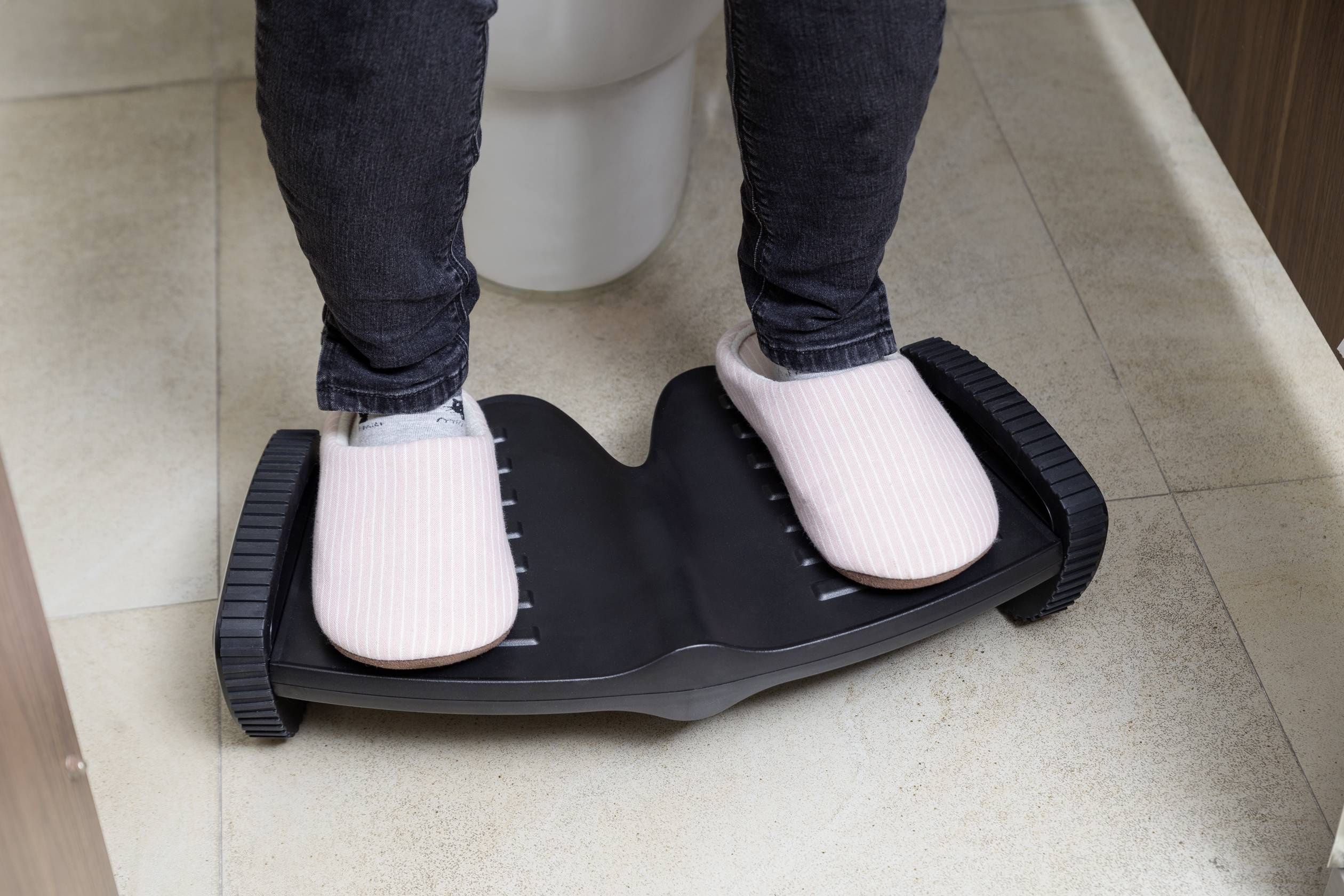 Eine Person steht auf einer schwarzen Balance-Plattform, trägt rosa Hausschuhe. Die Plattform hat gerillte Seiten und ist auf Fliesenboden.