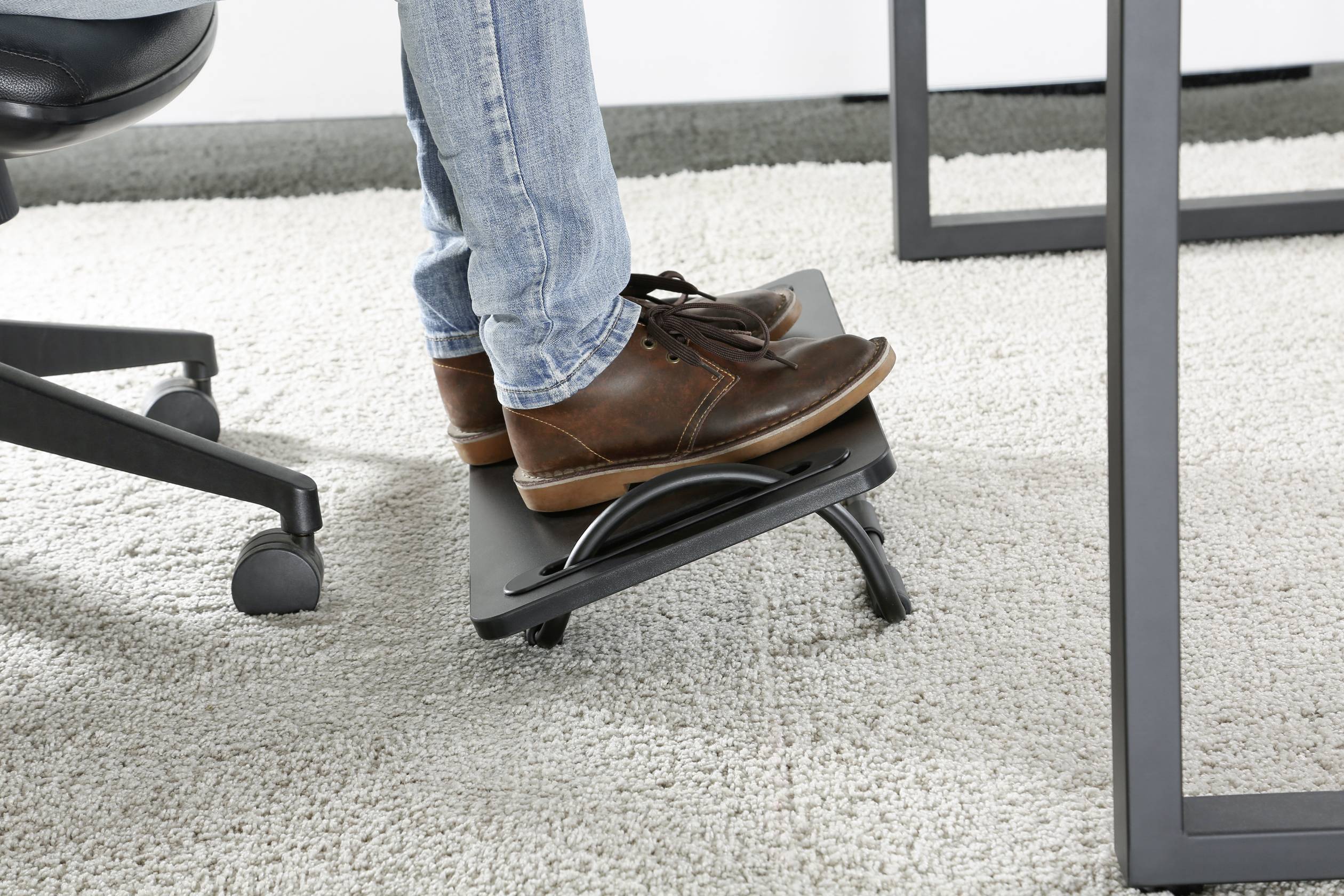 Person trägt Lederstiefel und verwendet eine schwarze Fußstütze unter einem Tisch, um die Füße anzuheben. Auf Teppichboden.
