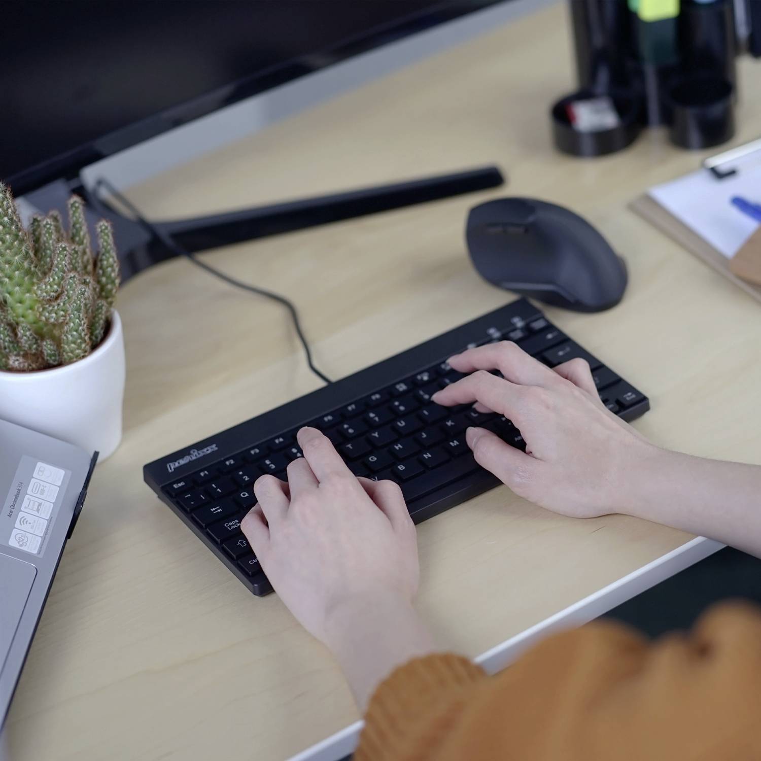 Eine Person tippt auf einer schwarzen Tastatur auf einem Holztisch, daneben eine Pflanze, Maus, Notizblock und Bürobedarf.
