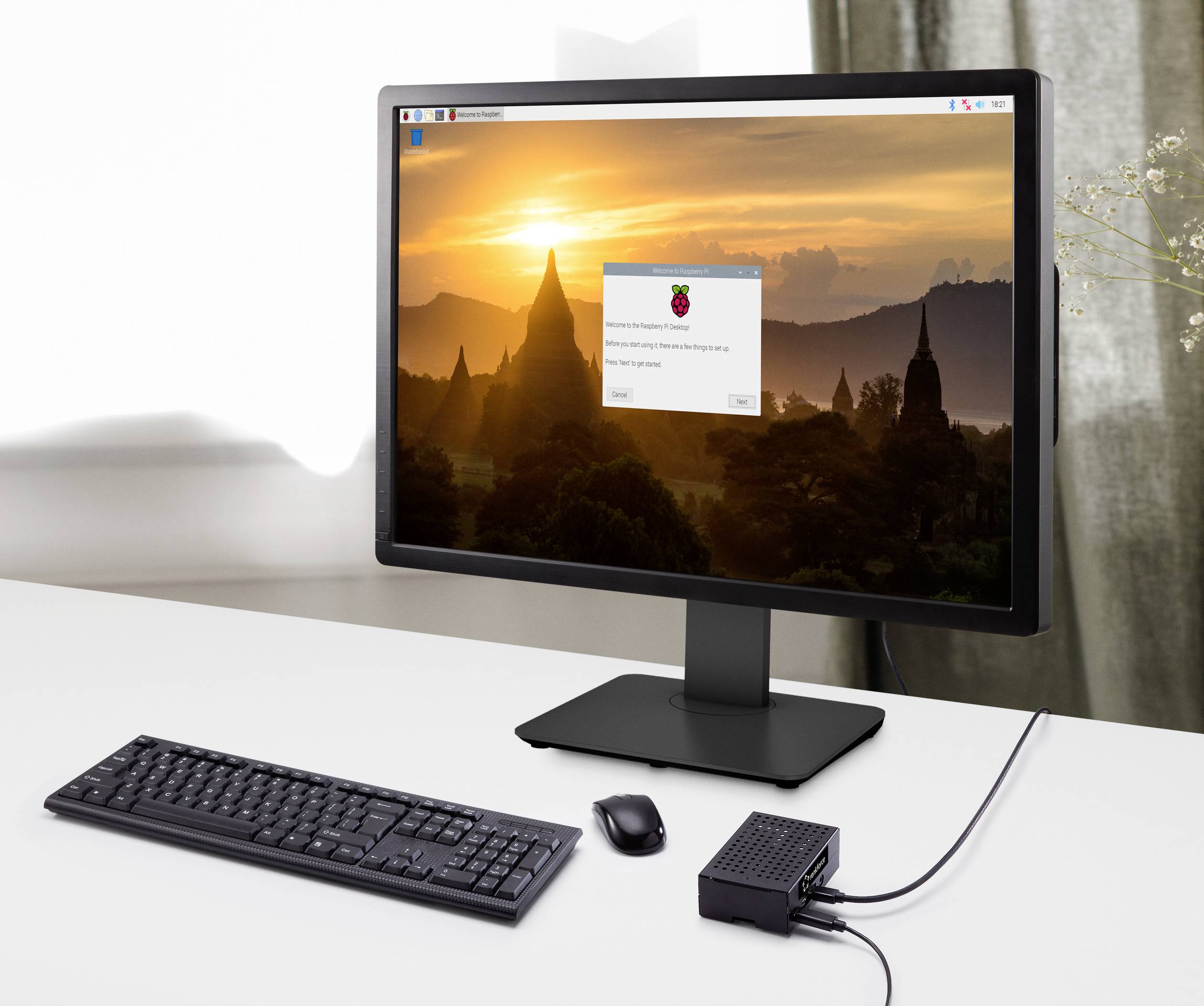 Computerbildschirm mit Landschaft und Raspberry Pi-Betriebssystemlogo, Tastatur, Maus und Raspberry Pi-Gerät auf weißem Tisch.