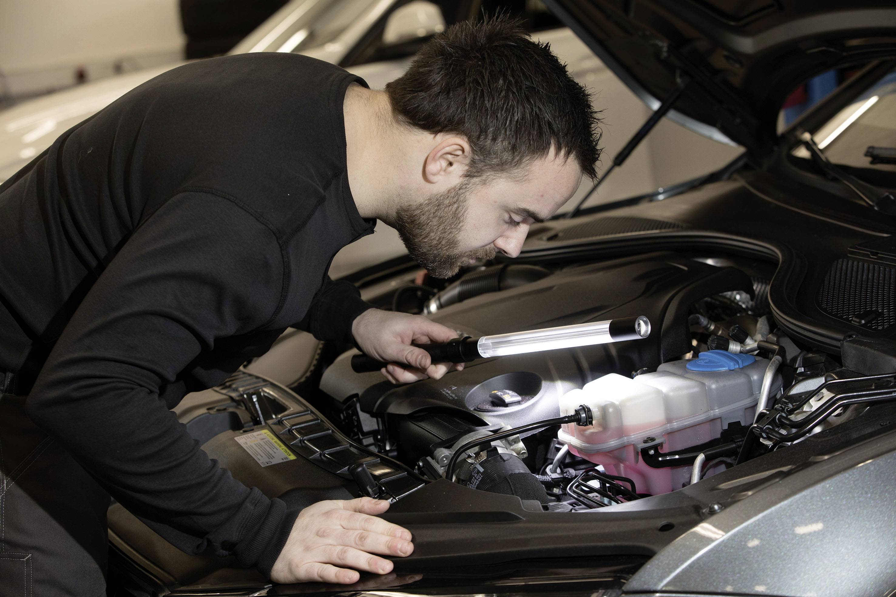 Ein Mechaniker inspiziert den Motorraum eines Autos mit einer Taschenlampe, um mögliche Probleme zu identifizieren.