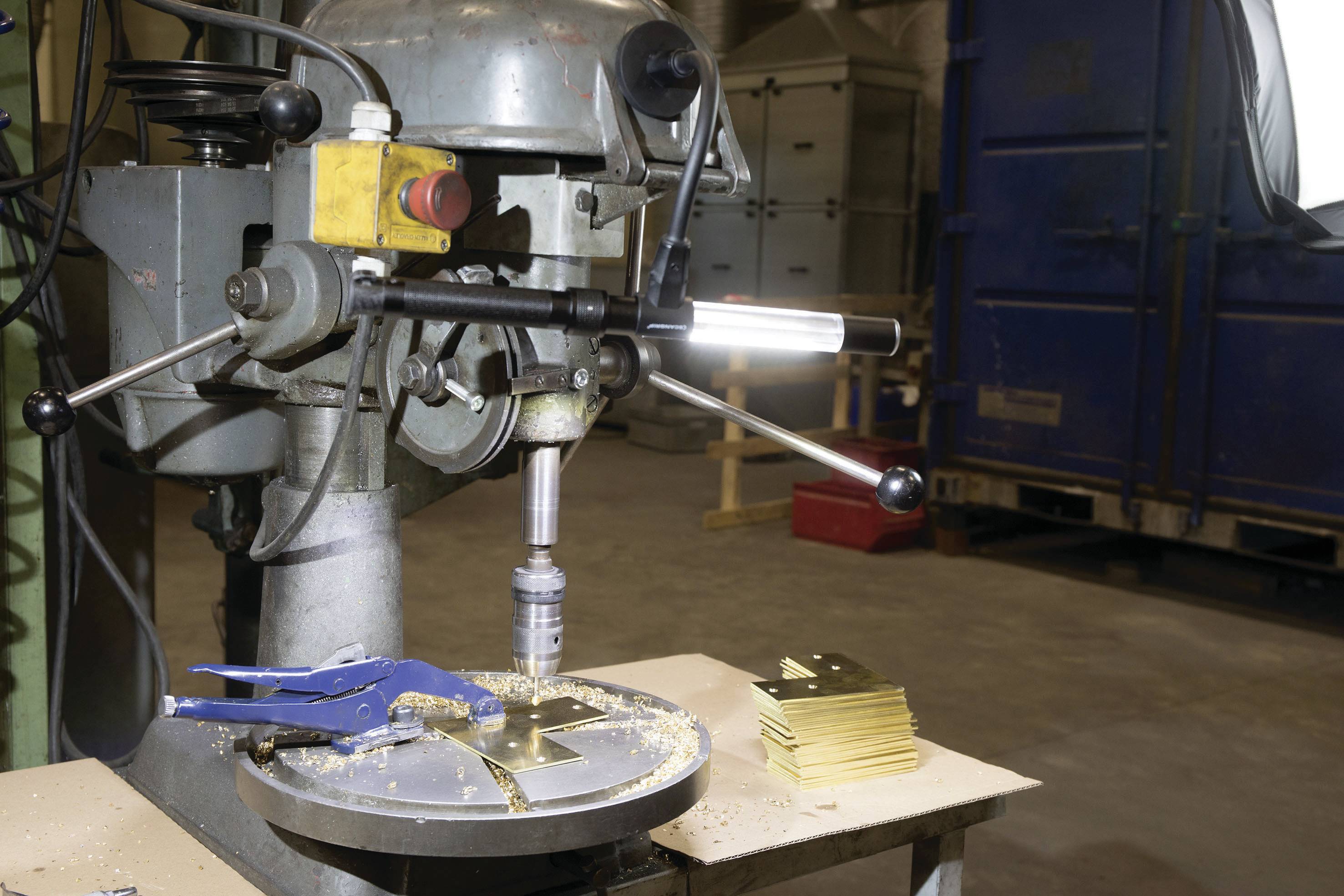 Eine Werkstatt zeigt eine Bohrmaschine mit eingespanntem Bohrer. Auf dem Tisch liegen gebohrte Metallplatten, umgeben von Spänen.
