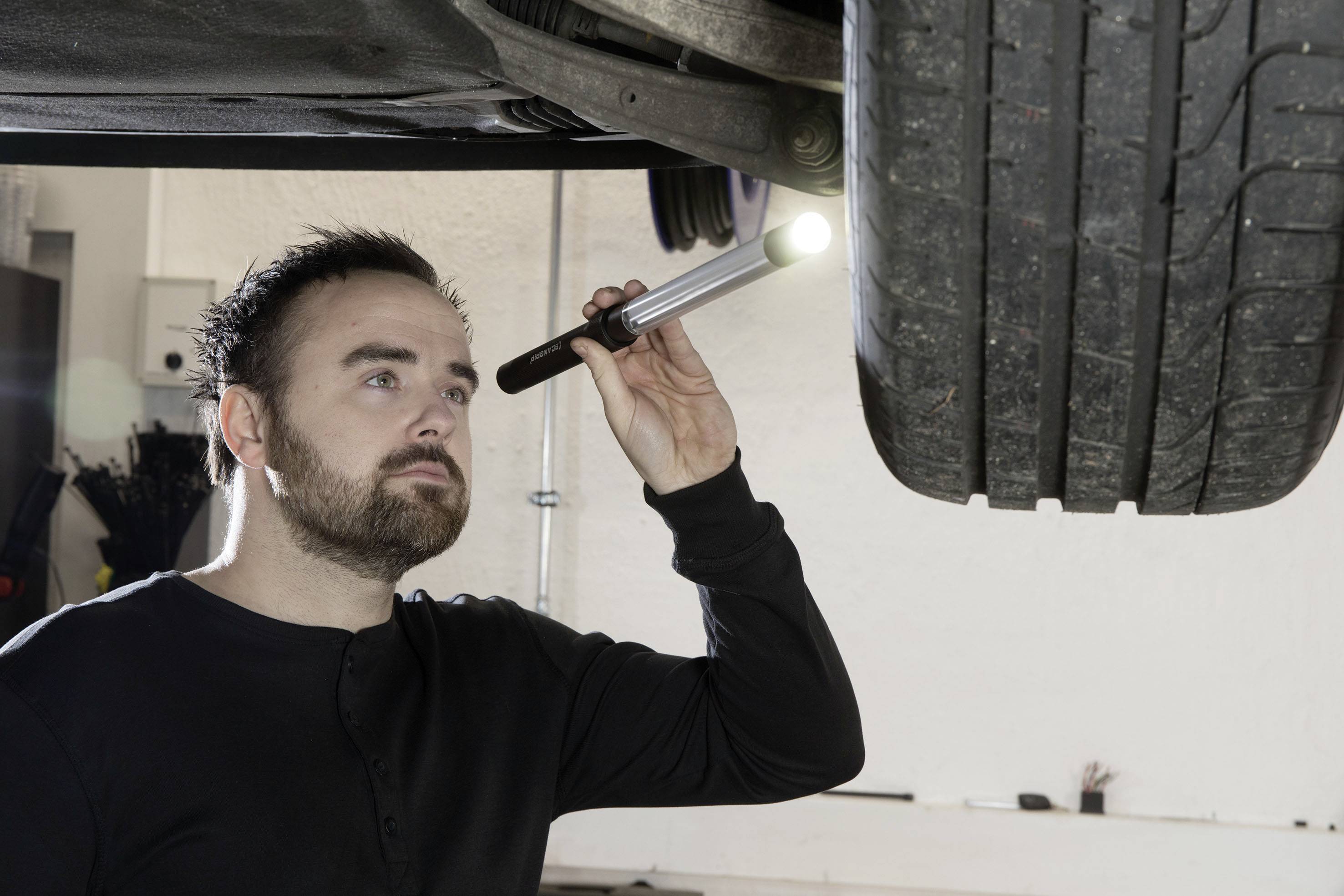 Eine Person inspiziert mit einer Taschenlampe den Unterboden eines angehobenen Autos in einer Werkstatt.