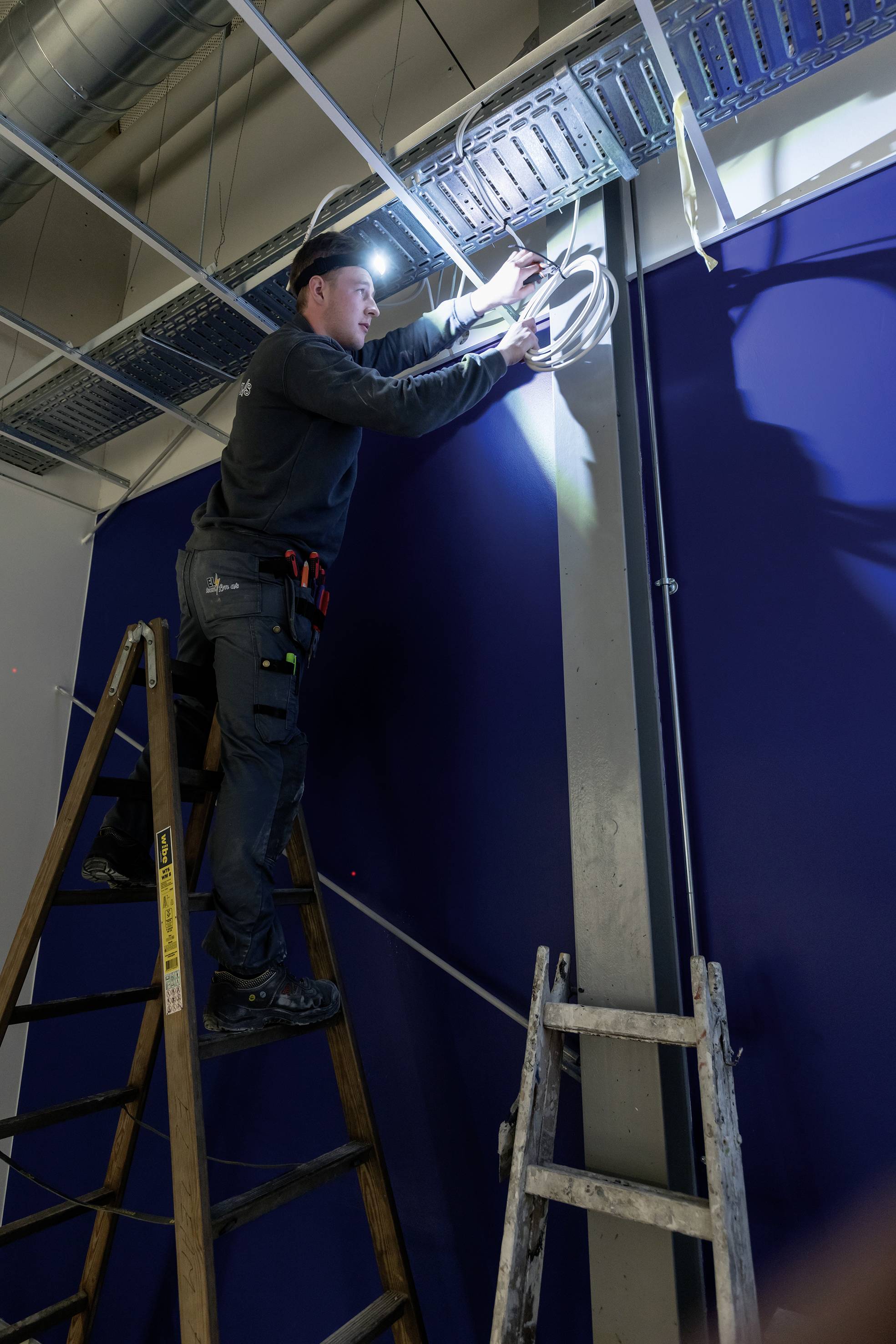Mann steht auf einer Leiter und installiert Kabel an der Decke in einem Raum mit blauen Wänden.
