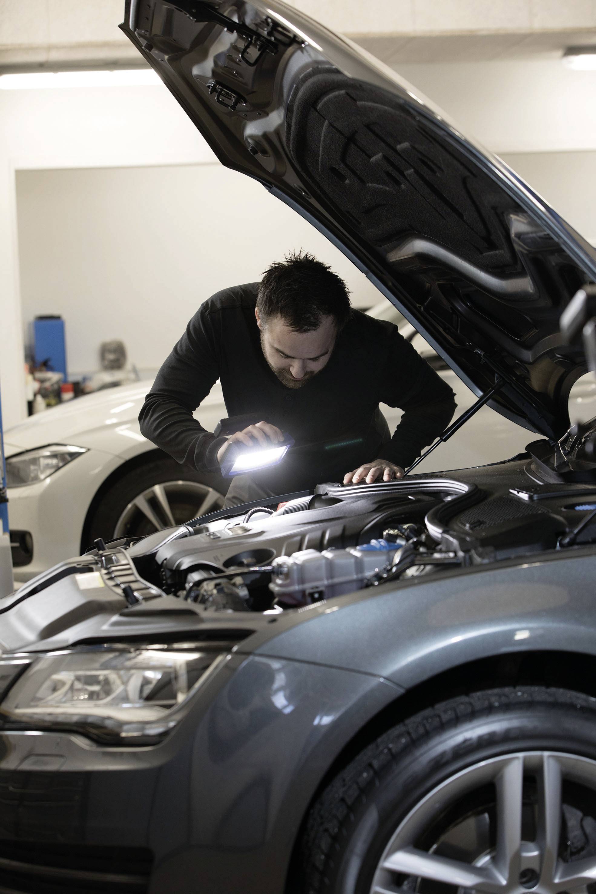 Mann inspiziert Motor eines Autos mit Taschenlampe in Werkstatt. Weitere Autos im Hintergrund.