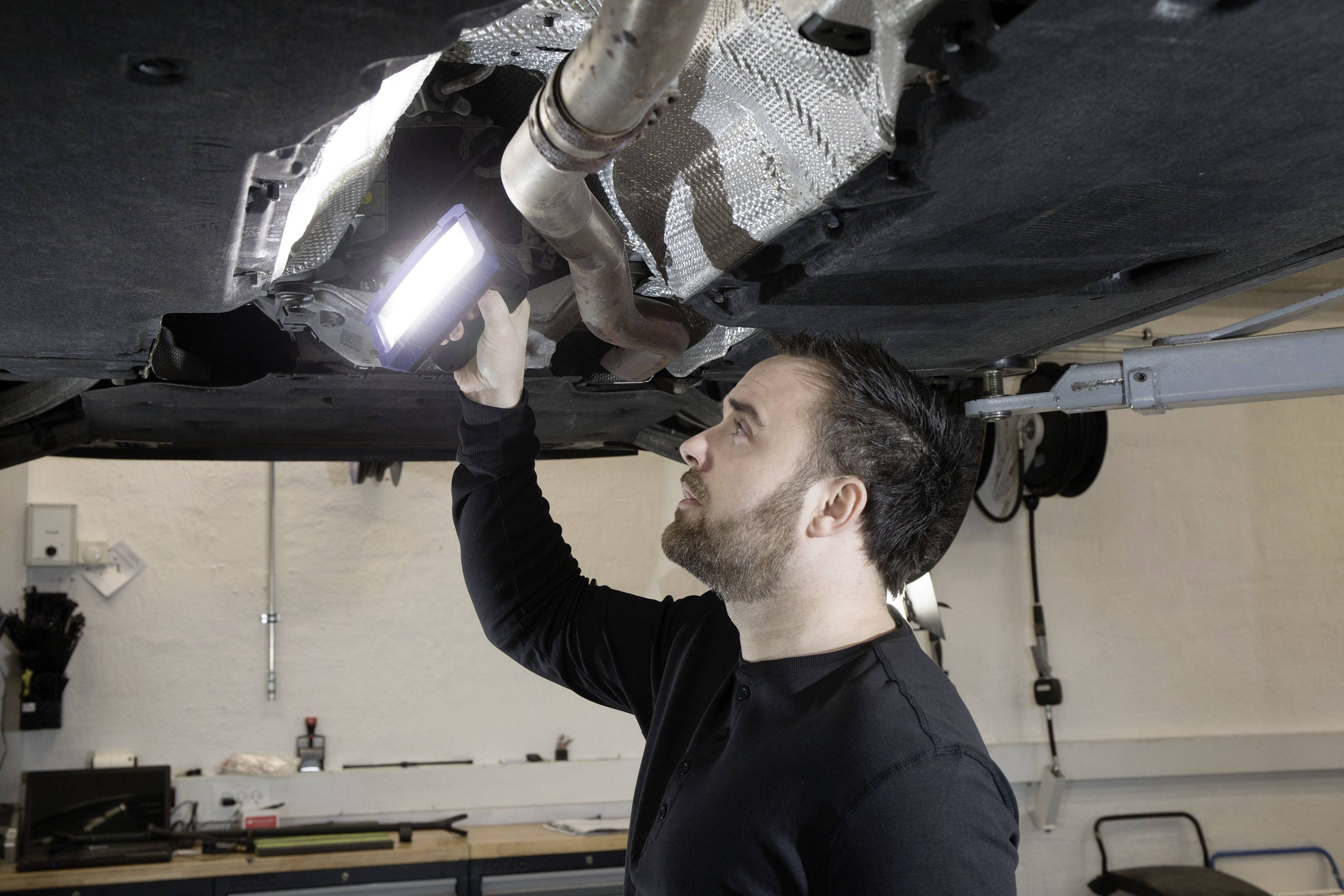 Ein Mann inspiziert den Unterboden eines Fahrzeugs mit einer leuchtenden Werkstattlampe in einer Werkstatt.
