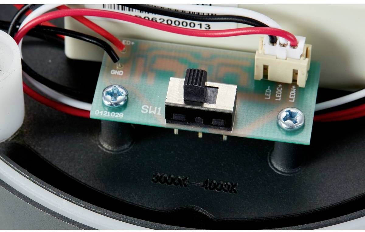 'Ein kleiner elektrischer Schalter auf einer Leiterplatte, umgeben von verschiedenen Kabeln und mit zwei Befestigungsschrauben befestigt.'