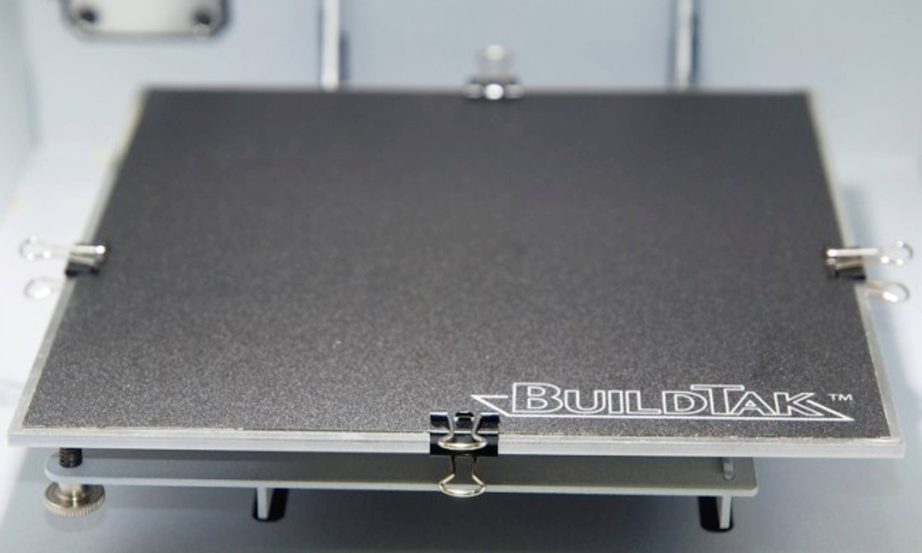 Buildtak-Oberfläche auf einem 3D-Druckerbett, bereit für den Druck. Die Oberfläche zeigt eine glatte, strukturierte Platte mit Clips.