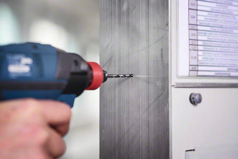 'Eine Hand hält einen Bohrer, der ein Loch in eine Metallwand bohrt. Rechts ist ein Plandokument sichtbar.'