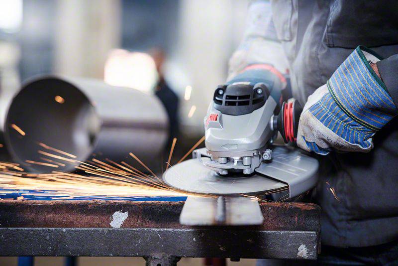 Ein Arbeiter trägt Handschuhe und benutzt einen Winkelschleifer, um Metall zu bearbeiten. Funken fliegen von der Metalloberfläche.