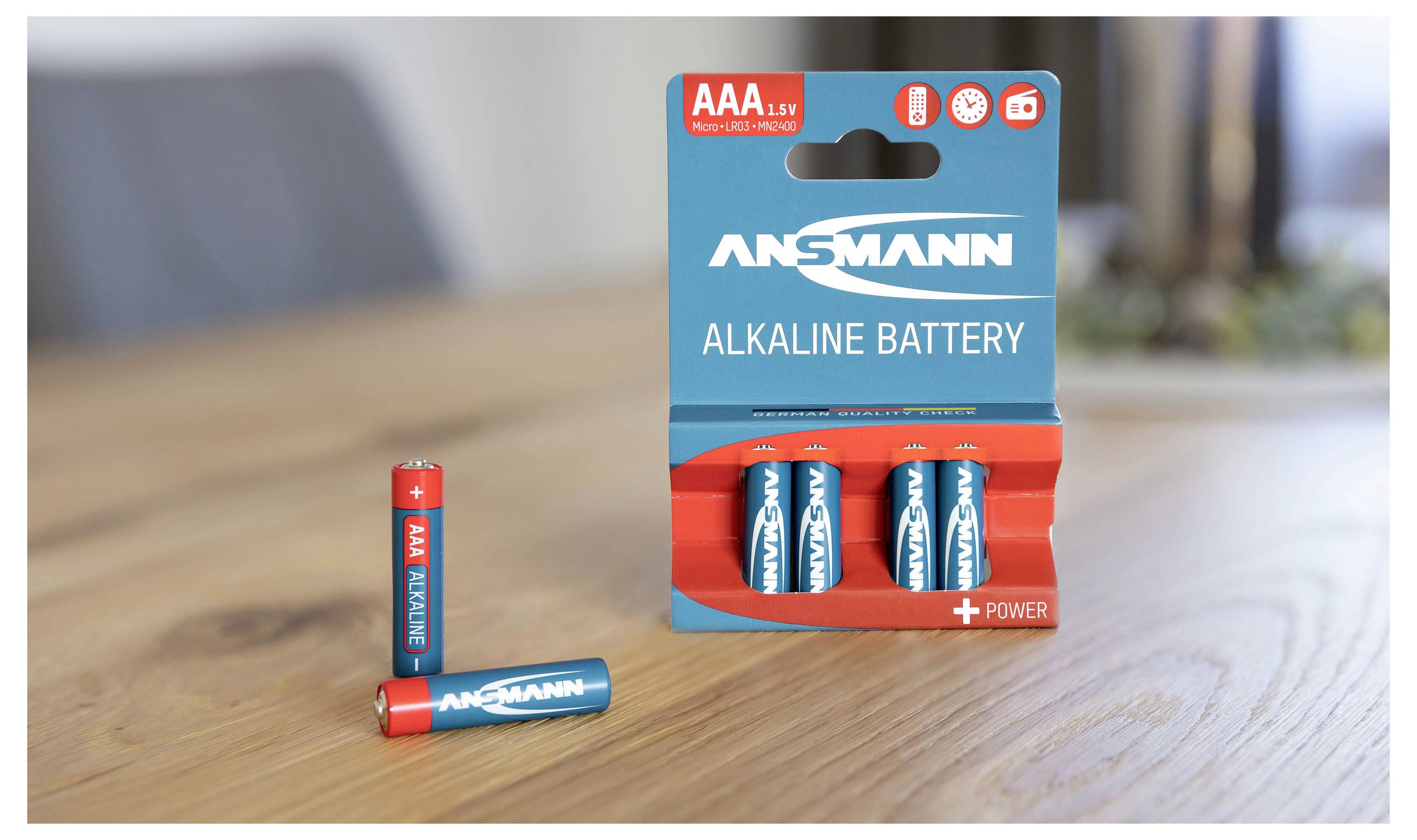Eine Packung mit drei blauen Alkaline-Batterien der Marke Ansmann, eine Batterie steht separat daneben auf einem Holztisch.